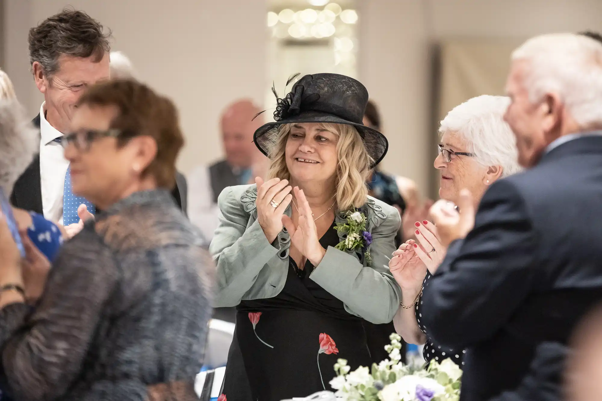 Group of people clapping at an indoor event. A woman in the center wearing a black hat and floral dress appears to be the focus.