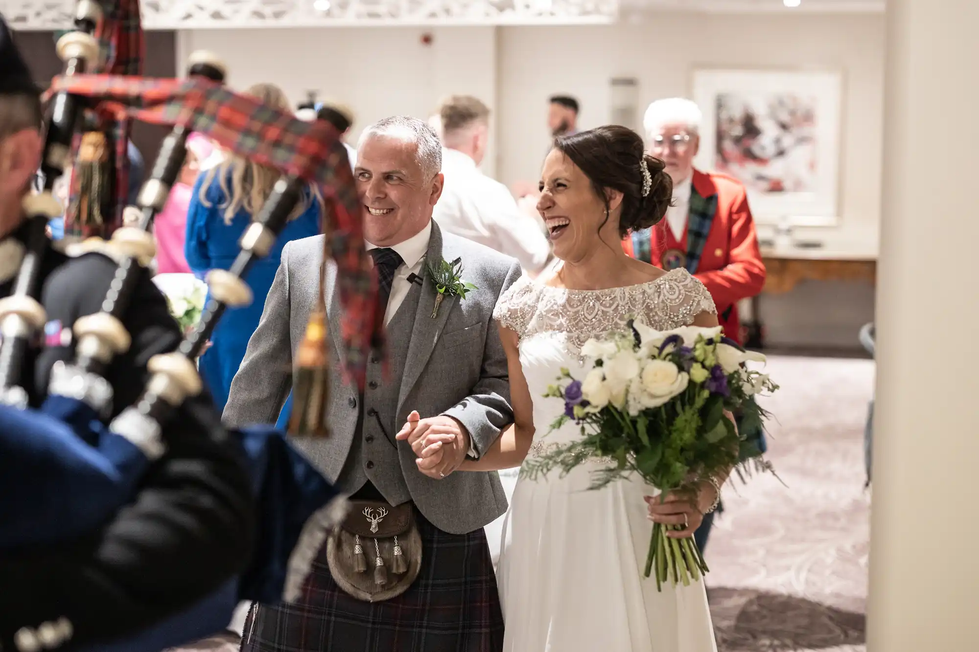 A couple, dressed in traditional attire, smile and hold hands while walking. The woman holds a bouquet and people, including a bagpiper, are in the background.