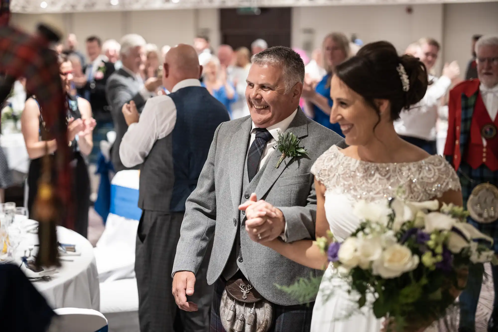 A bride and groom, both smiling, walk hand-in-hand through a crowd of applauding guests at a wedding reception. The groom is in a gray suit, and the bride is in a white dress holding a bouquet.