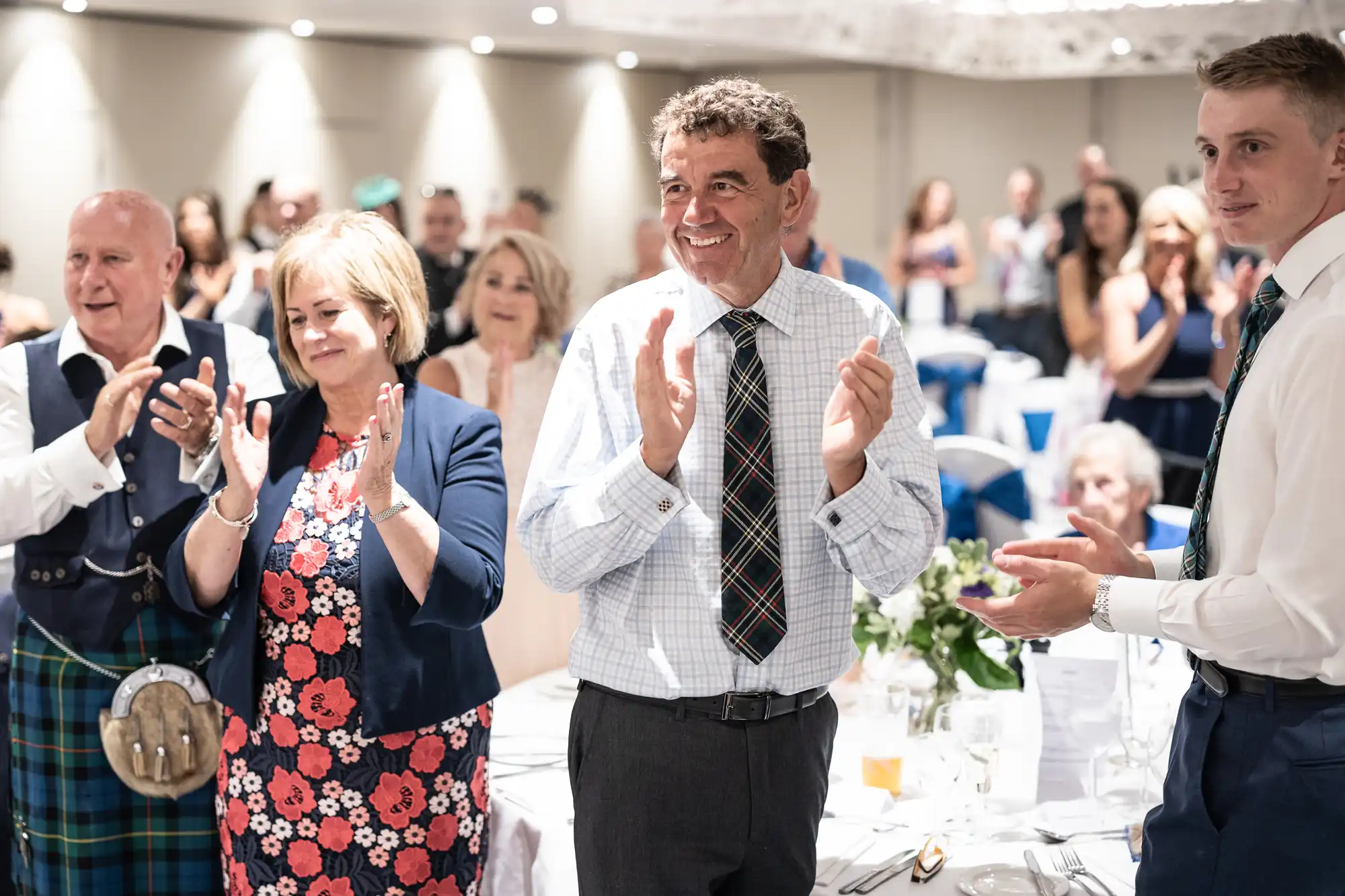 A group of people, some in formal Scottish attire, stand and applaud in a brightly lit room with white decor.