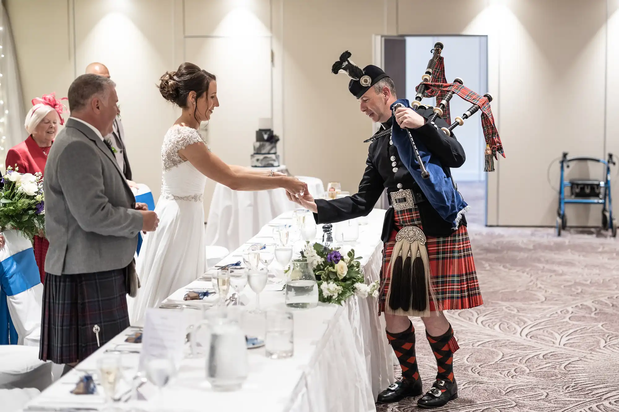 A bride shakes hands with a bagpiper dressed in traditional Scottish attire at a wedding reception, with guests looking on.