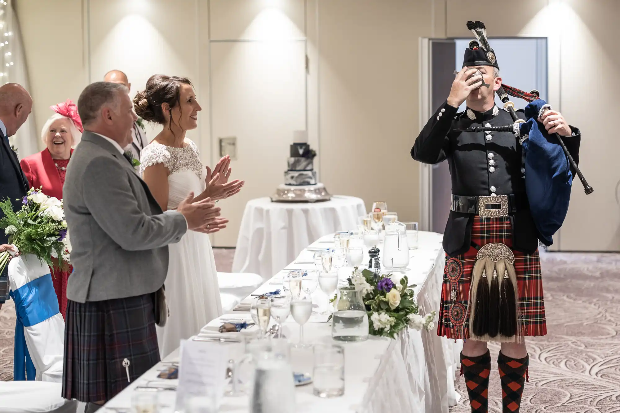 A bagpiper performs at a wedding reception while the bride and groom, dressed in wedding attire and traditional Scottish kilts, applaud. The head table is set with white linens and floral arrangements.