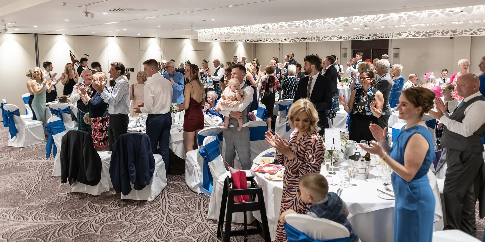 A group of people wearing formal attire stand and applaud at a large banquet-style event with round tables and blue-accented chair covers.