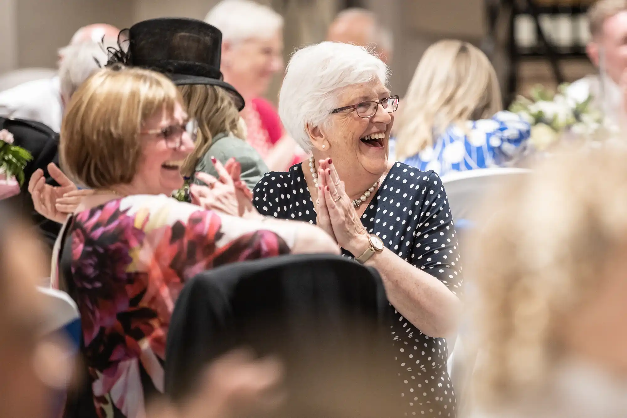 Two elderly women, one in a floral dress and one in a polka dot dress, smiling and clapping at a social gathering. Other attendees are visible in the blurred background.