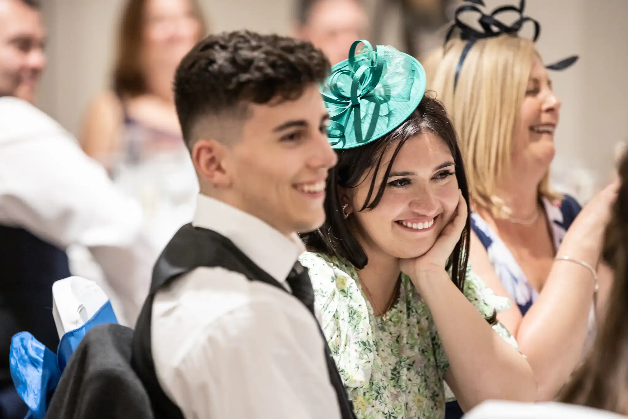 Two people, a young man and a young woman, sit smiling at an event. The woman wears a green fascinator, and others are visible in the background, also smiling.