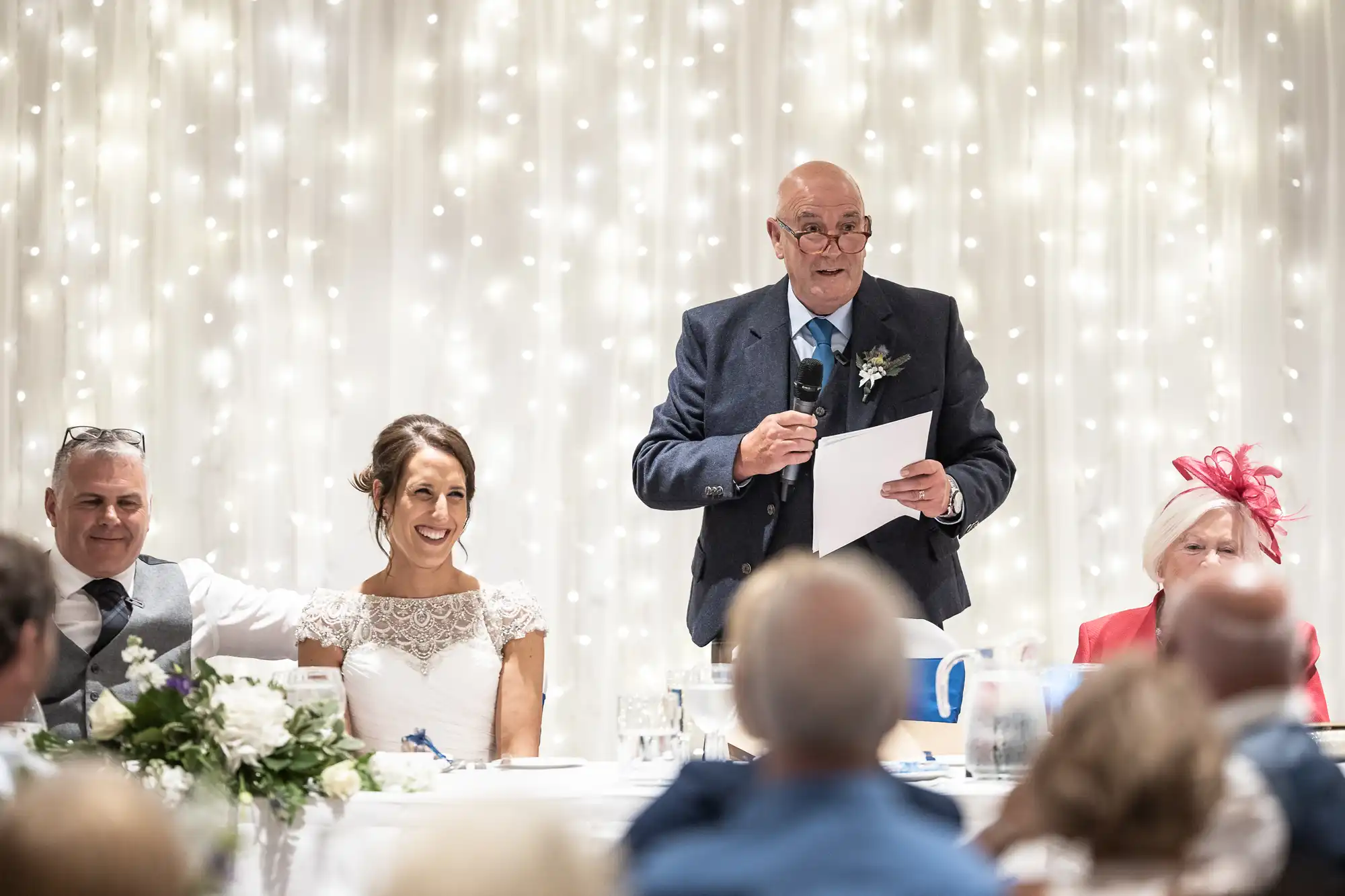 An older man is giving a speech behind a table at a wedding, with a bride, groom, and elderly woman seated beside him under twinkling lights.