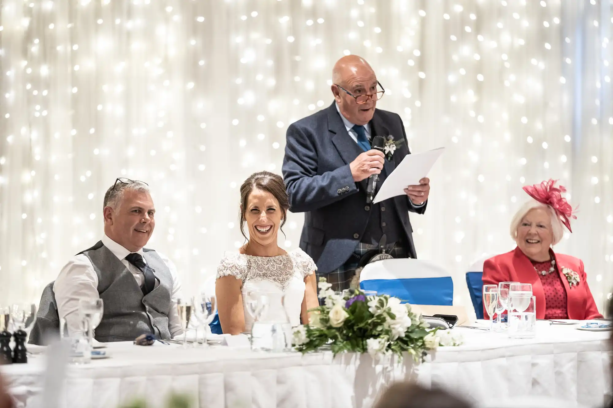 A man in a suit is giving a speech at a wedding reception. The bride, groom, and an older woman are seated at the head table, smiling. The background features string lights.