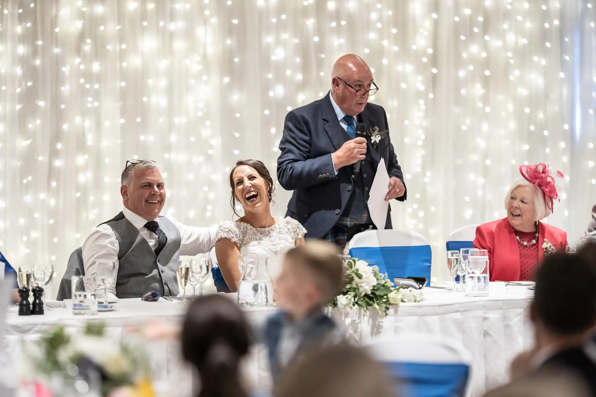 A man in a suit gives a speech with a microphone while a bride and groom sit and laugh at a table decorated with flowers and glasses. A woman in a pink outfit and hat smiles sitting nearby.