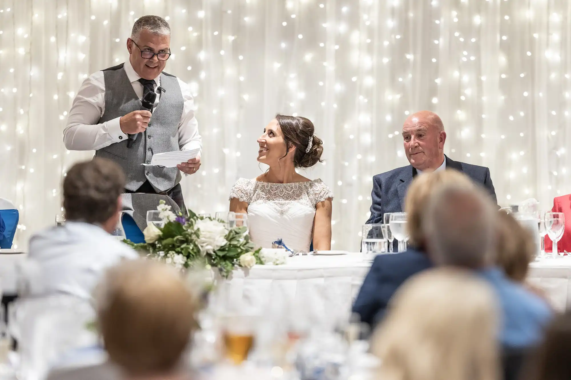 A man stands and speaks into a microphone while holding a paper, next to a bride and another man, during a wedding reception with twinkling lights in the background.