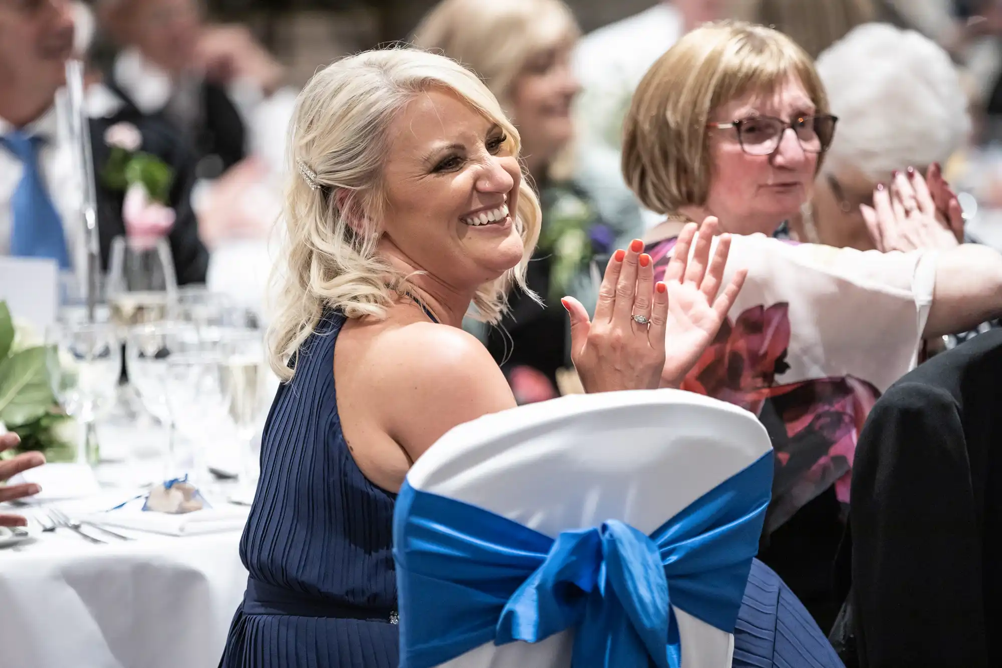 A woman with blonde hair and a blue dress is smiling and clapping at an event. She is seated at a decorated table with other attendees in the background.