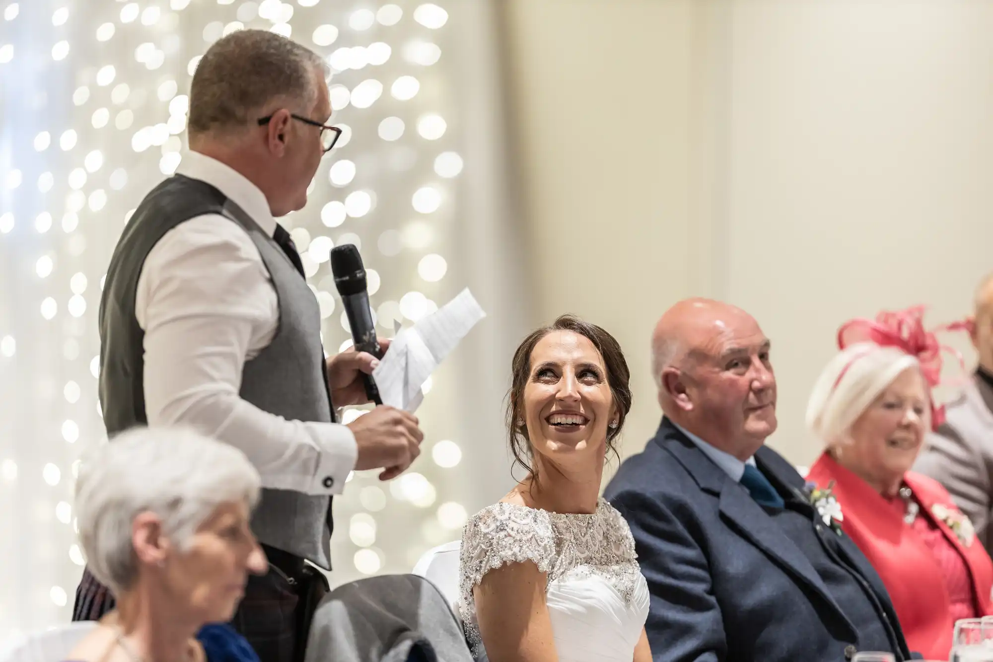 A man in a vest speaks into a microphone while a smiling woman in a white dress looks at him. Several seated individuals are in the background, dressed in formal attire.