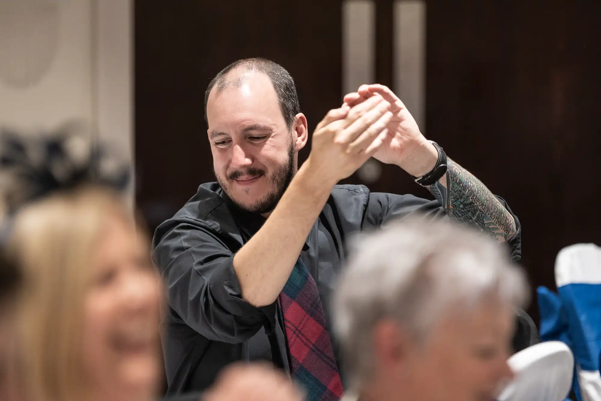 A man with a beard and tattoos, wearing a black shirt and checkered tie, smiling and clapping in a room with blurred people in the foreground.