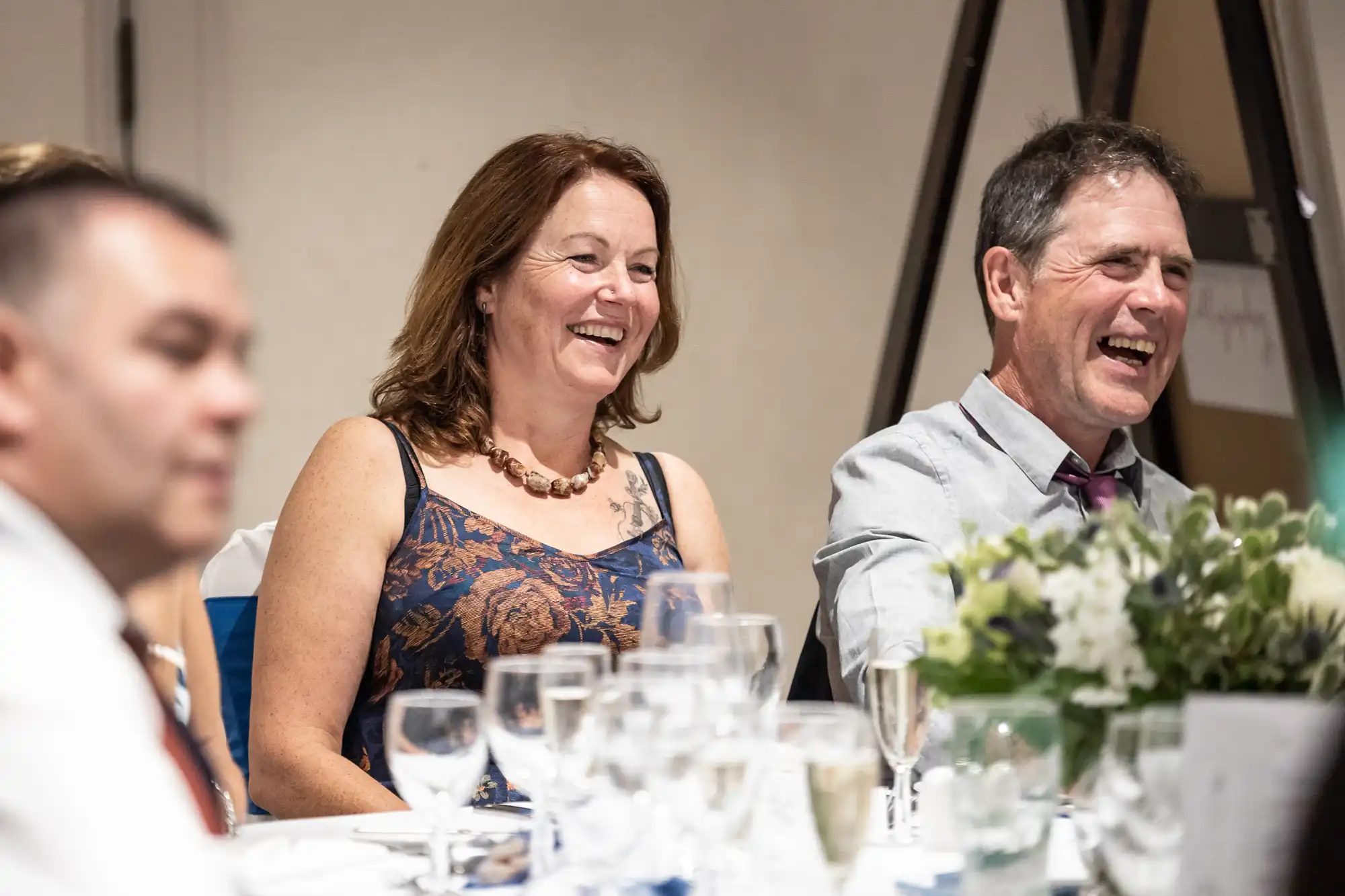 A woman and a man are sitting at a dining table, smiling and laughing. Empty wine glasses and a flower arrangement are on the table in front of them.