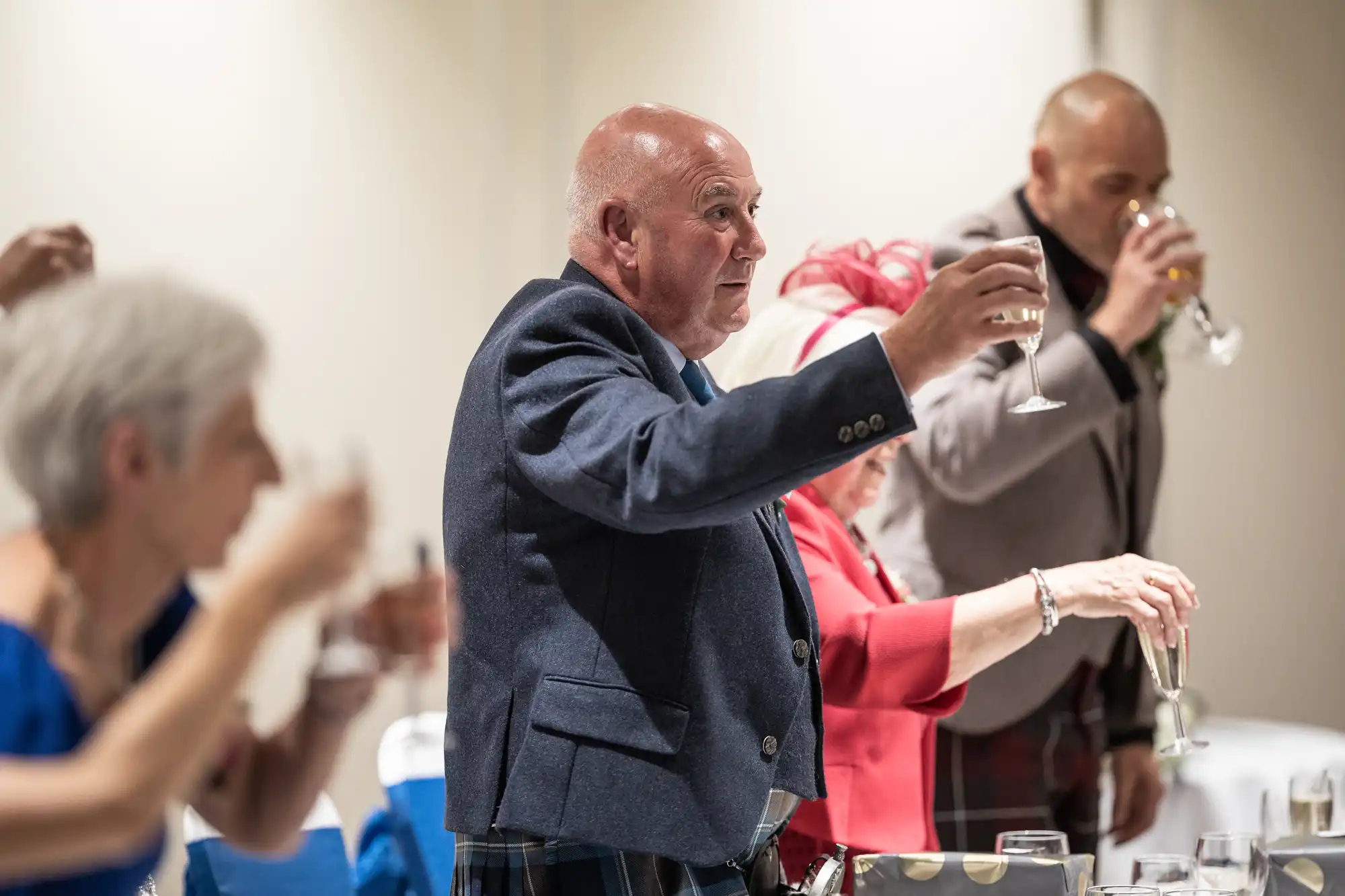 Several people in formal attire raise their glasses in a toast. The focus is on a man in a blue jacket holding a wine glass.