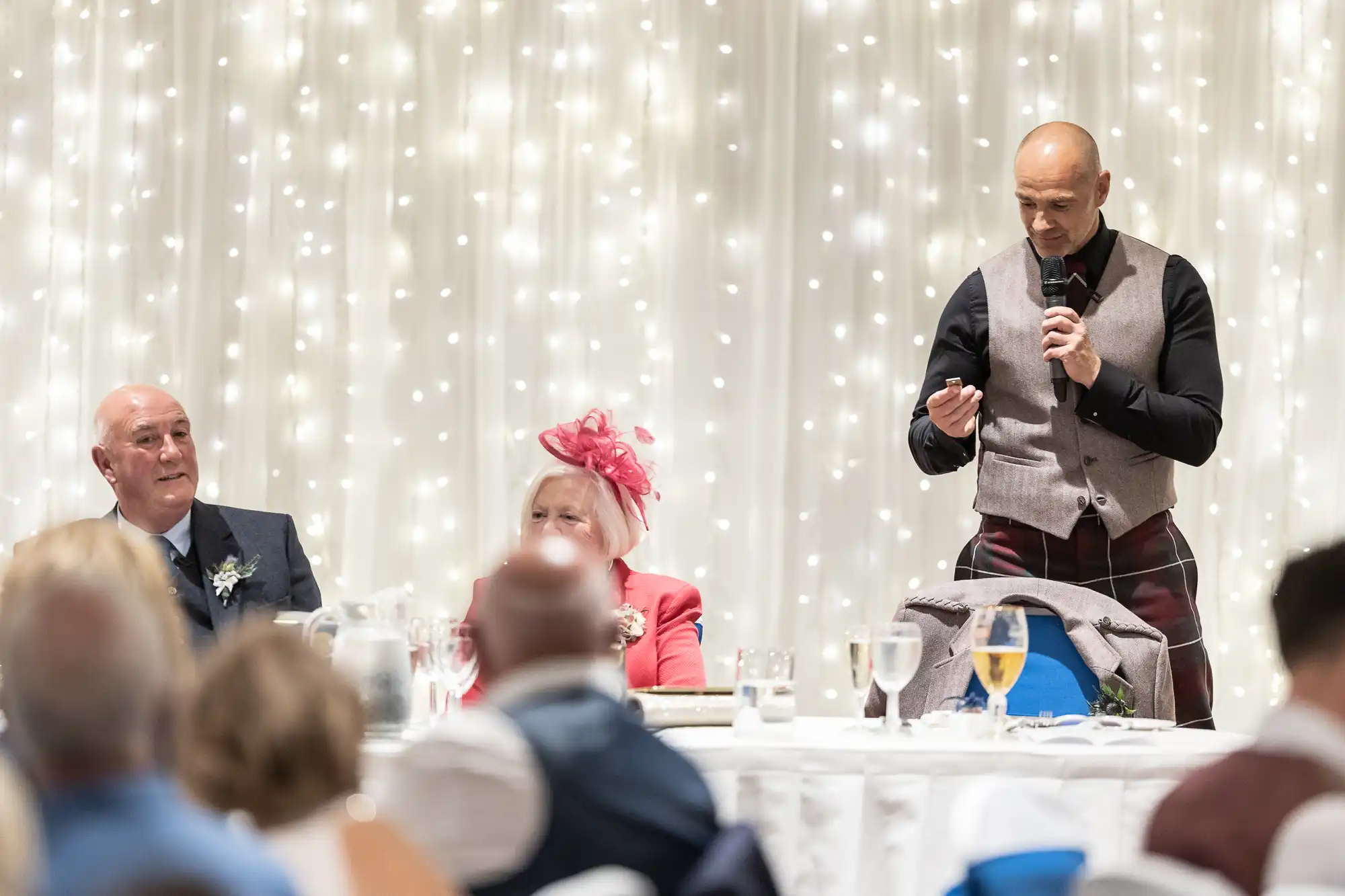 A man standing and speaking into a microphone; two seated individuals, one wearing a pink hat, listen attentively in a room decorated with string lights.