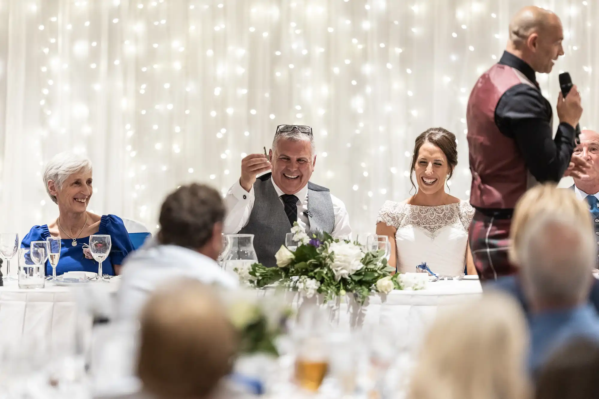 A man in a tartan vest gives a speech at a wedding reception. The bride and groom, seated at a long table with guests, listen and smile. White string lights hang in the background.