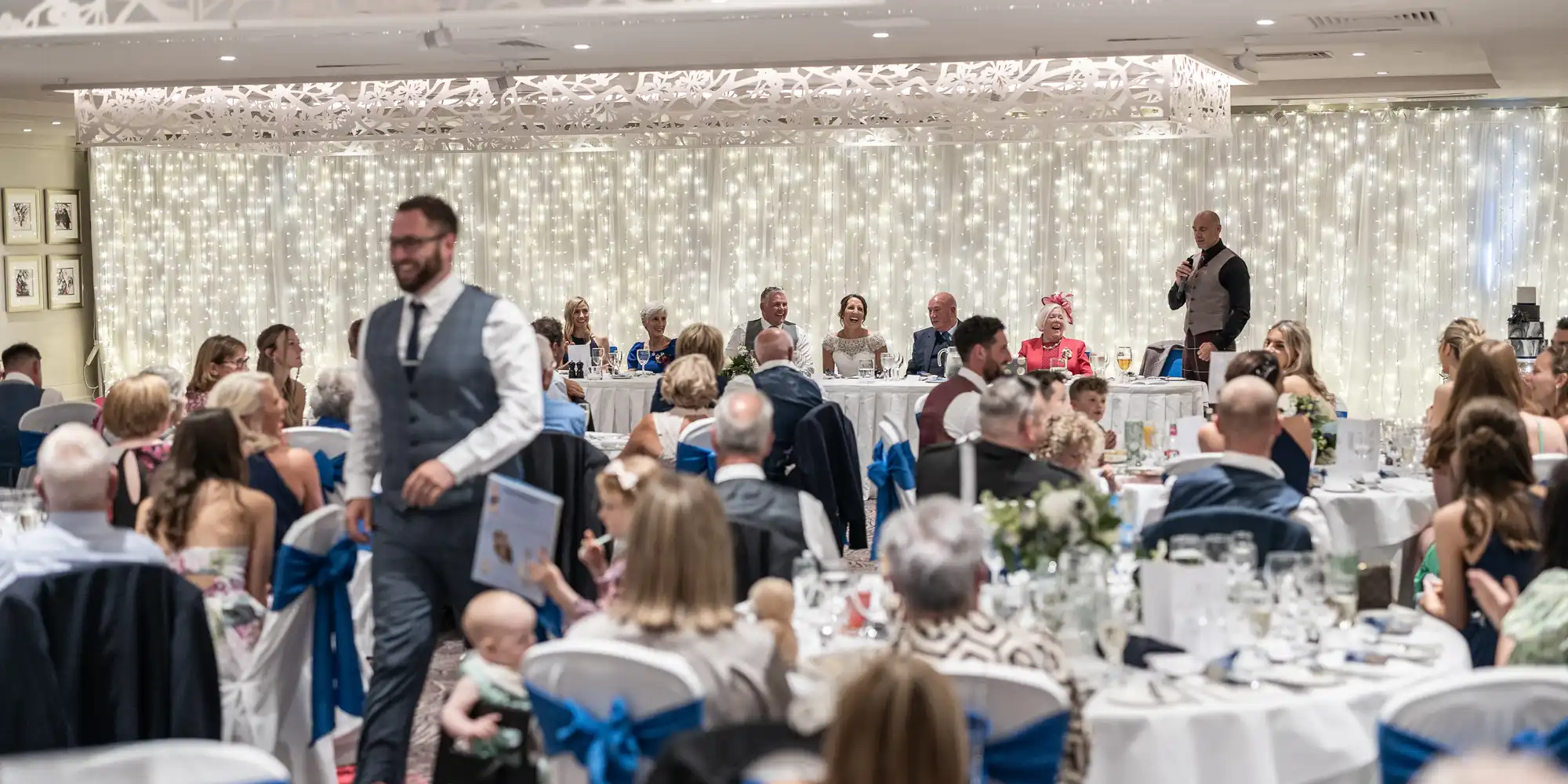 A formal event gathering with people seated at tables and a head table in the background. One person is speaking at a podium while another walks through the room. The room is decorated with lights.