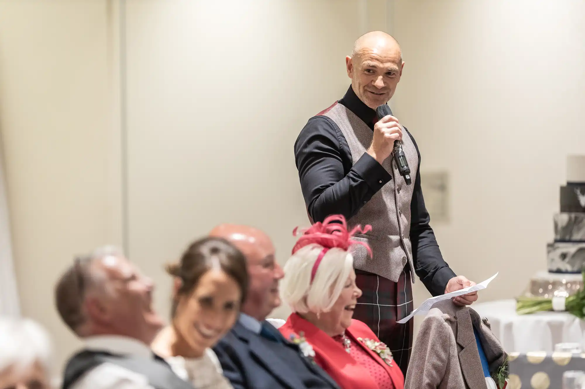 A man stands and speaks into a microphone while holding a piece of paper. Several seated people in the foreground are smiling and looking towards him.