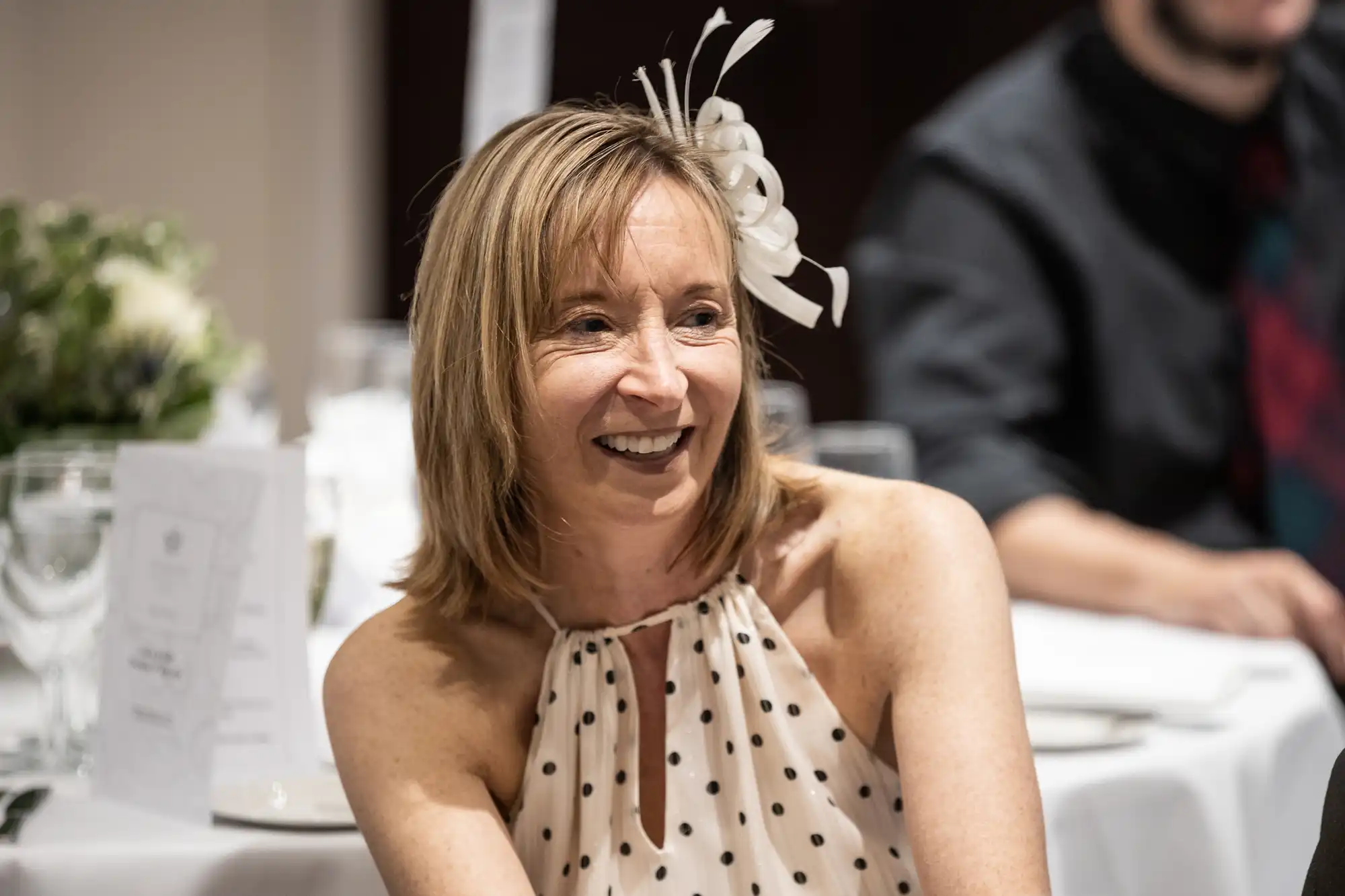 Woman with shoulder-length hair wearing a polka dot halter top and a white fascinator, smiling at an indoor event.