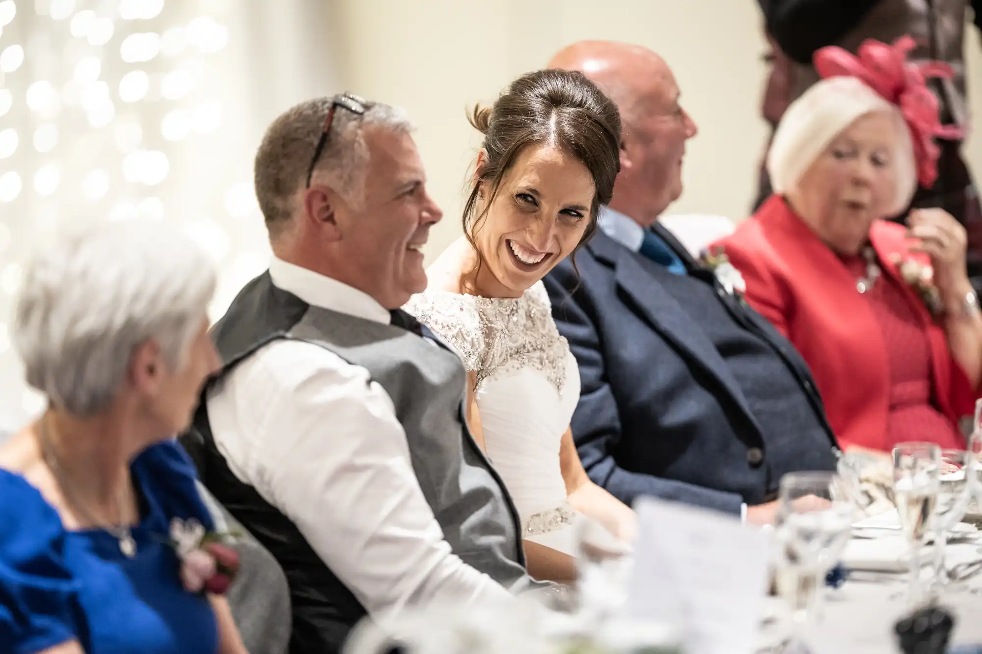 A woman in a wedding dress sits beside a man in a vest, smiling and looking at him. They are seated at a table with other guests dressed formally.