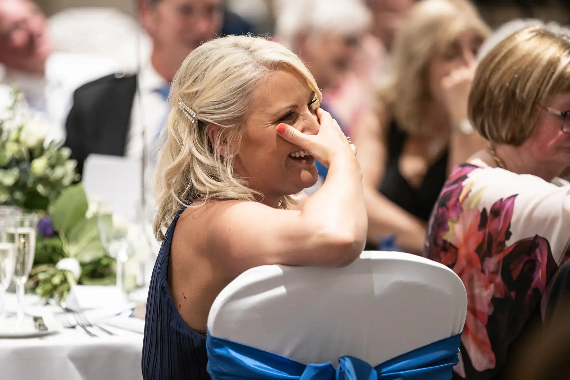 A woman with blonde hair, wearing a dark blue dress, laughs with her hand covering part of her face while sitting at a white table during an indoor event.