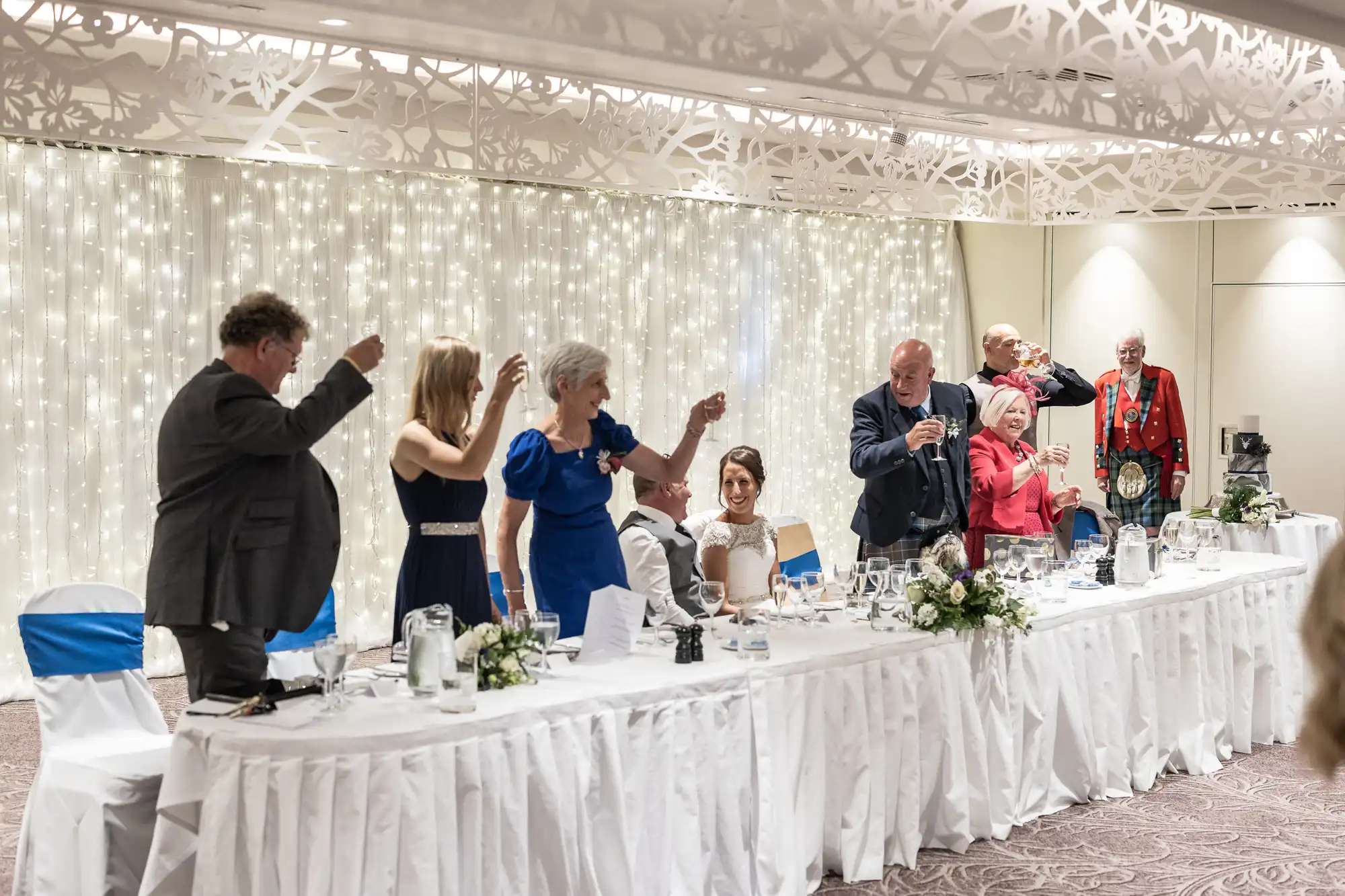 A group of people dressed in formal attire stand and raise glasses at a decorated head table during a celebration, with a backdrop of hanging string lights.