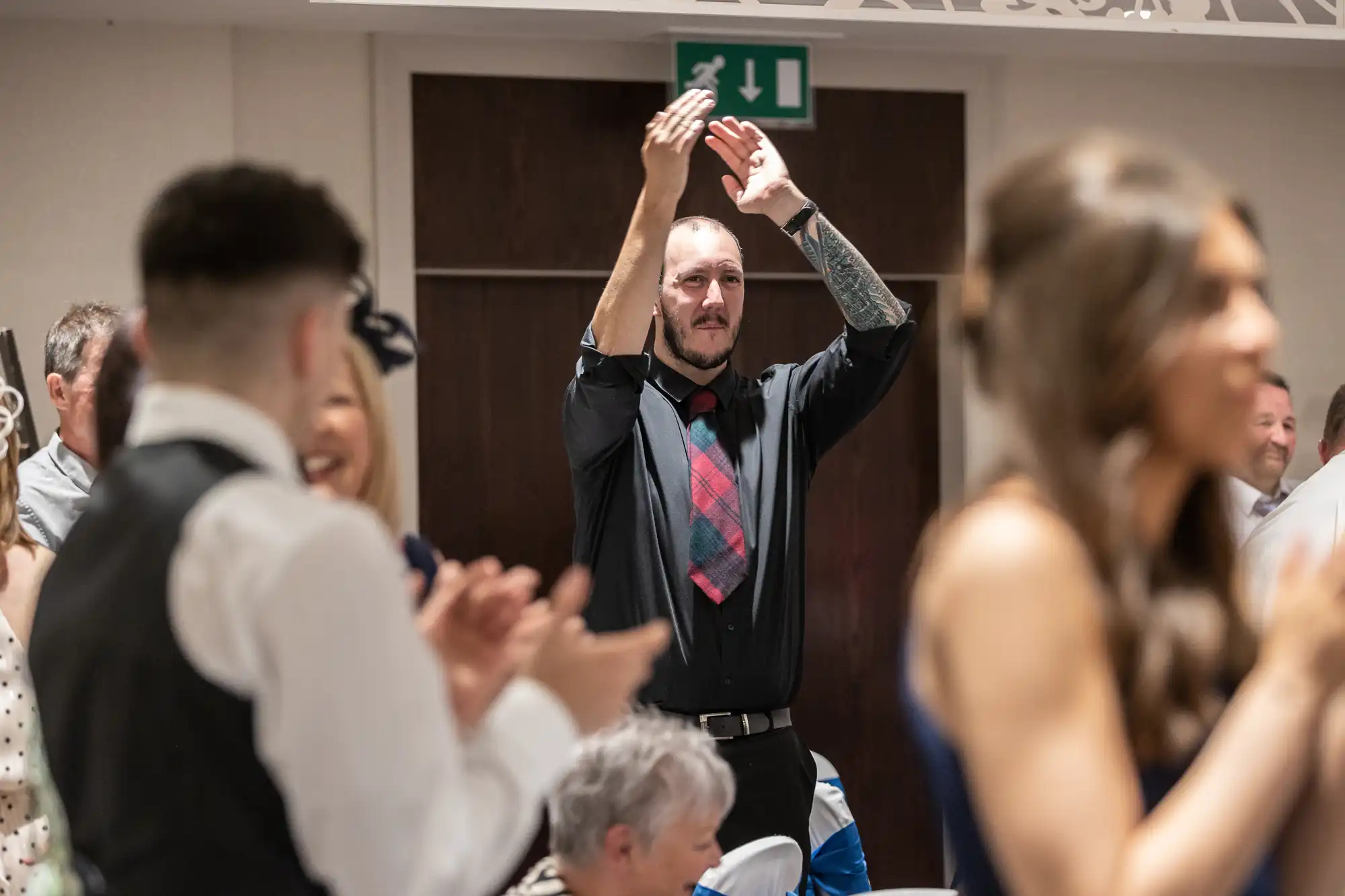 A man with tattoos, wearing a black shirt and red plaid tie, stands clapping at an indoor event. People around him are also clapping or looking in his direction.