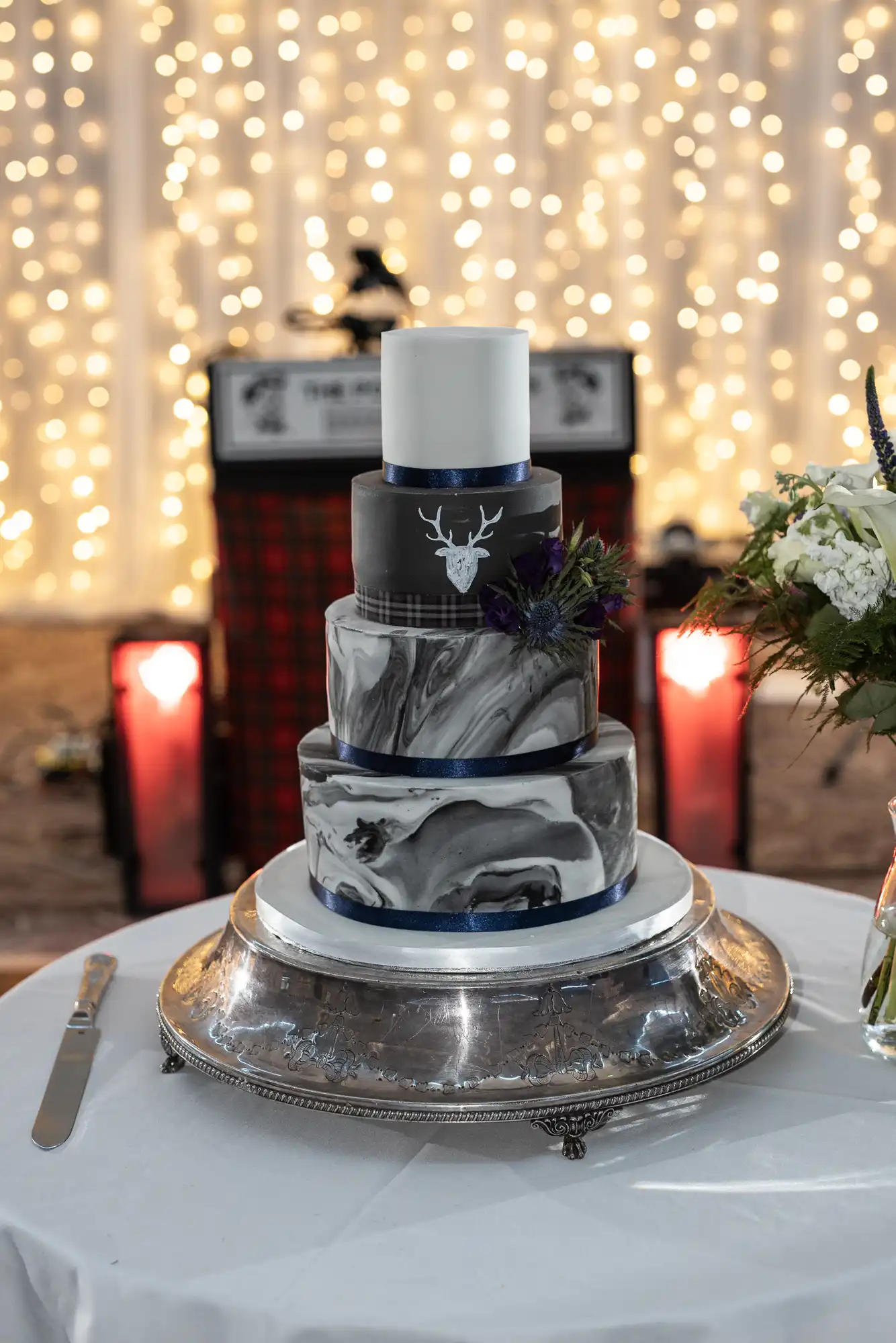 A four-tiered, black and white marbled wedding cake with a deer design on one tier sits on a silver stand. In the background are twinkling lights and a flower arrangement on the right.