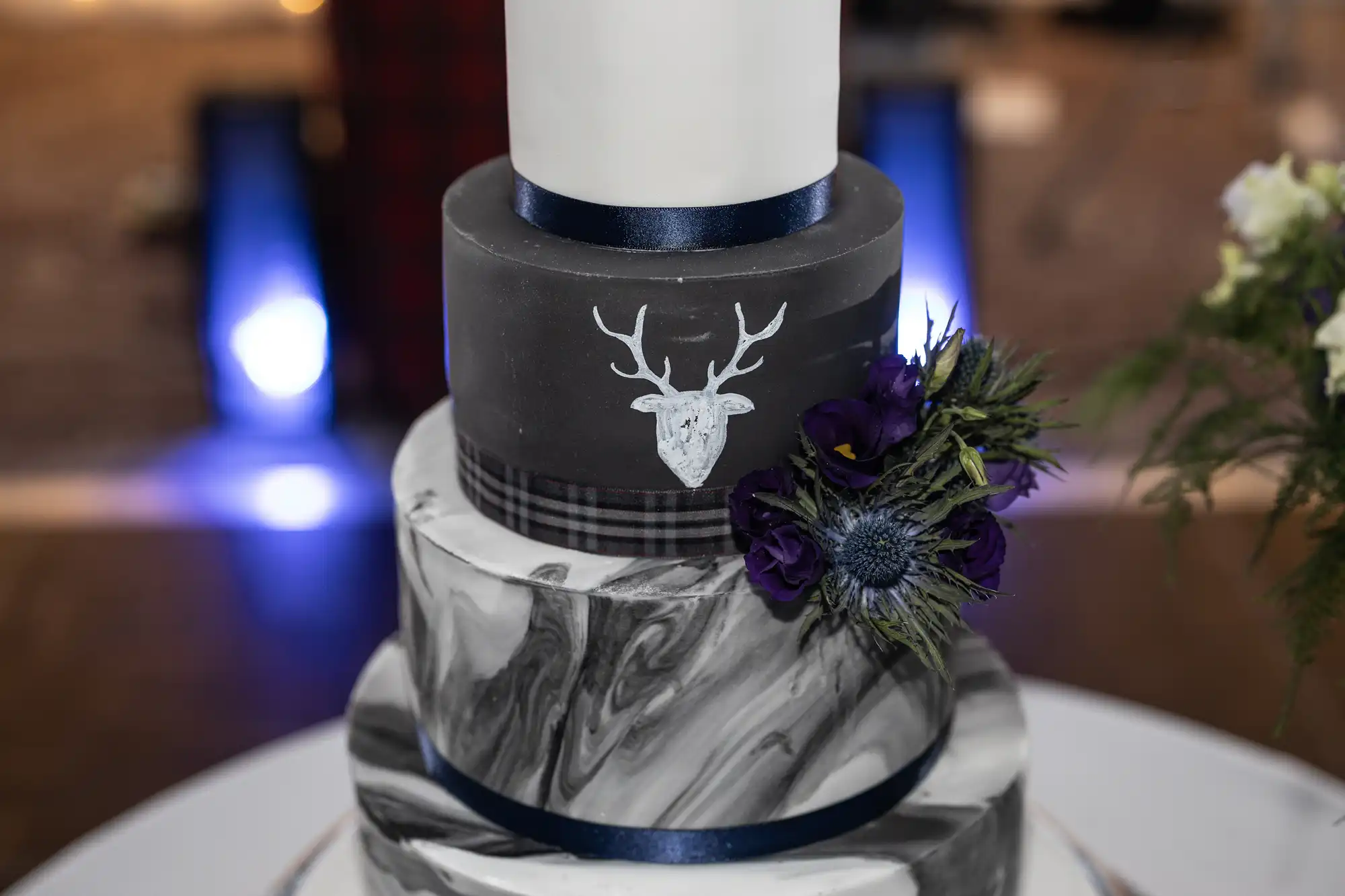 A three-tiered cake with a marble-patterned bottom tier, a gray middle tier featuring a white deer head design, and adorned with purple flowers and greenery.