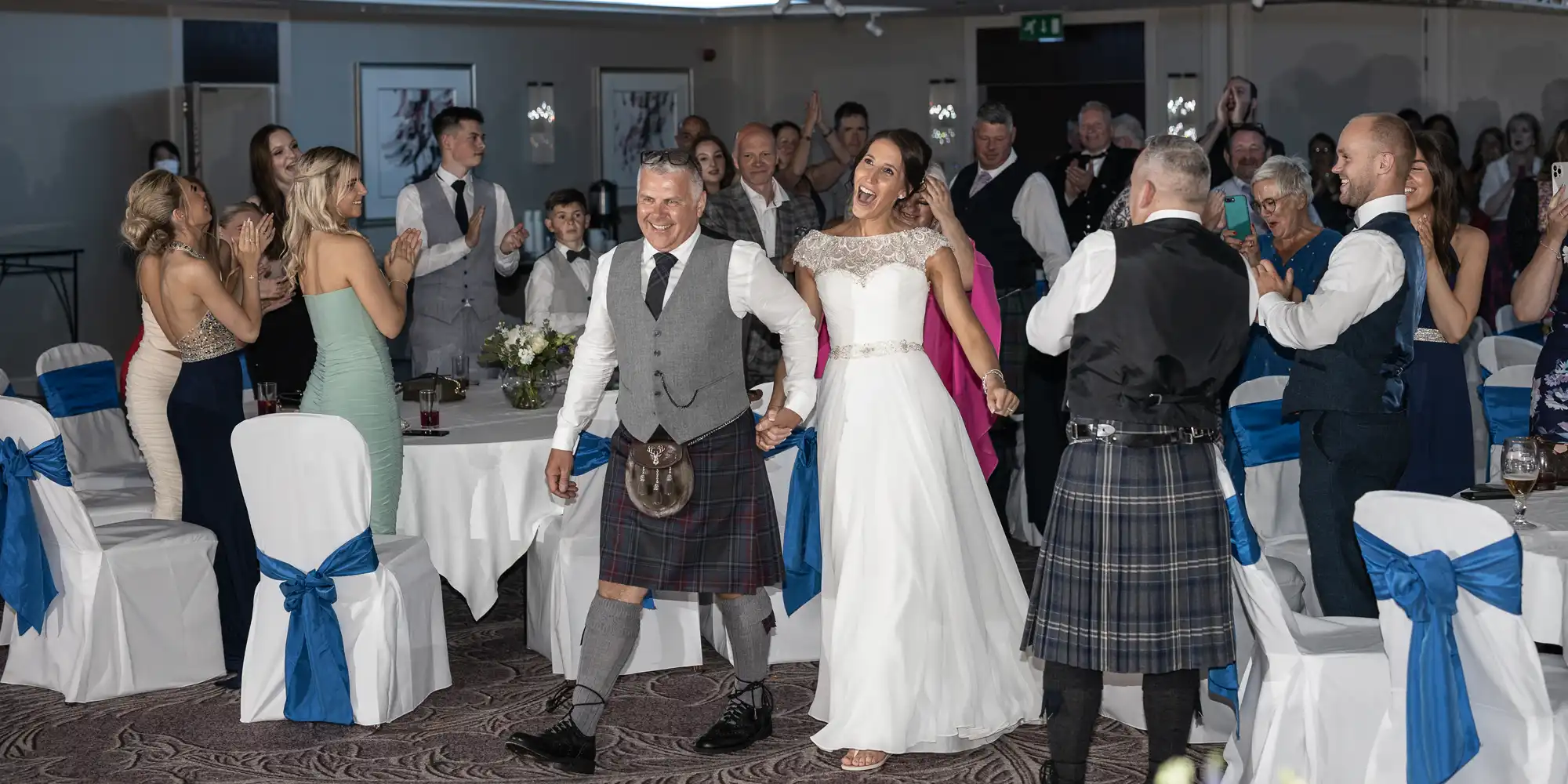 A bride and groom, dressed in traditional Scottish attire, walk hand in hand through a reception hall filled with applauding guests.