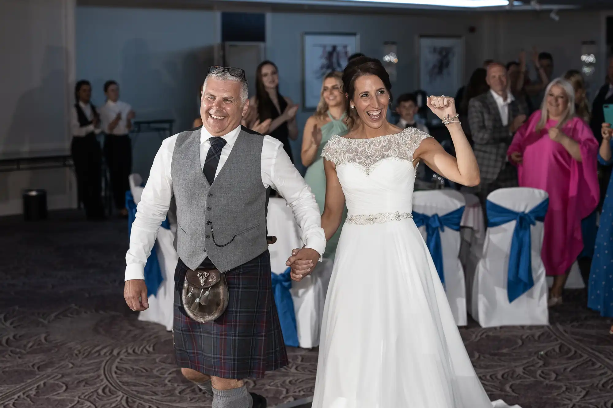 A bride and groom walk hand-in-hand, smiling and celebrating, while guests applaud. The groom wears a traditional kilt, and the bride wears a white wedding dress.