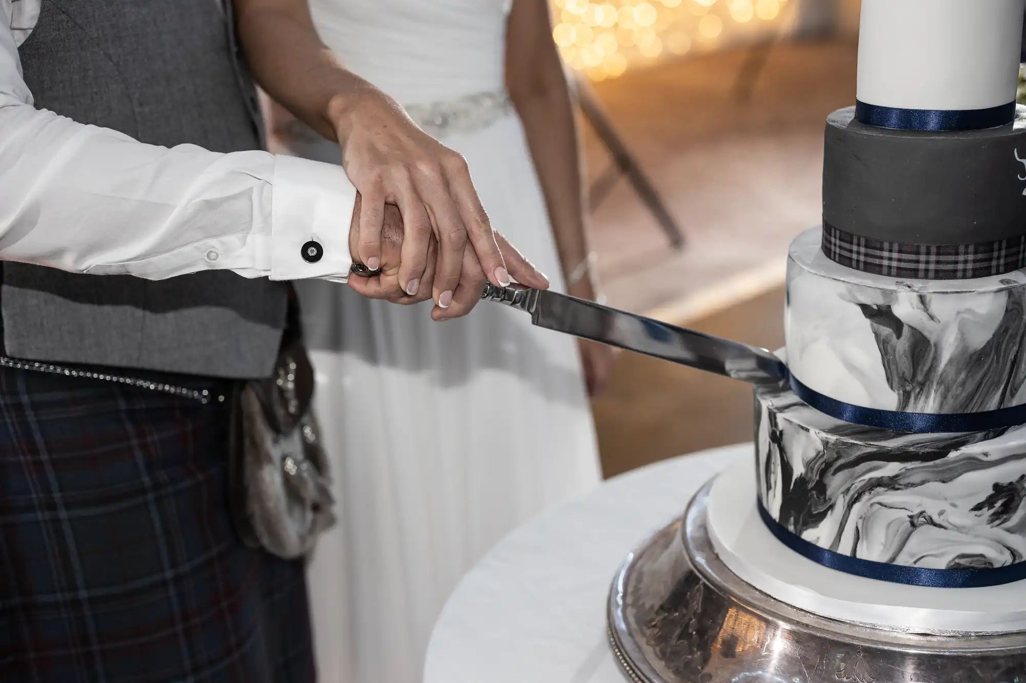 A couple slices a black-and-white marble-themed wedding cake together, their hands sharing a knife. The bride wears a white dress, and the groom appears to be in a vested outfit.