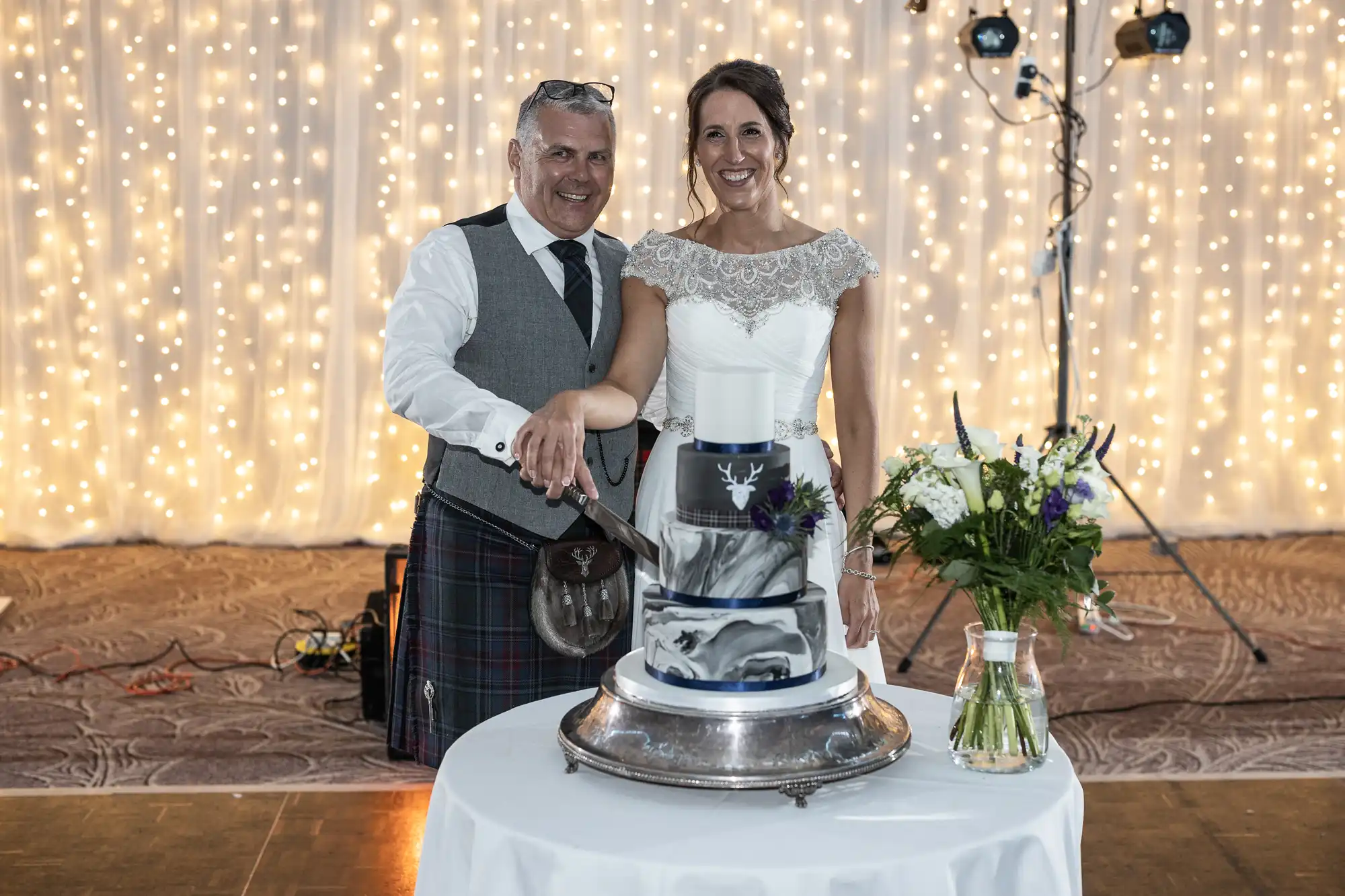 A couple, dressed in formal attire, smiles while cutting a wedding cake. The backdrop is decorated with twinkling lights and a bouquet stands on the table next to the cake.