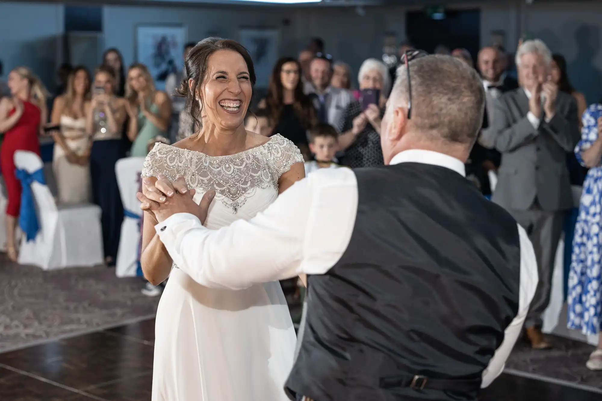 A bride in a white dress and an older man in formal attire dance joyfully together while a crowd of people watches and applauds in the background.