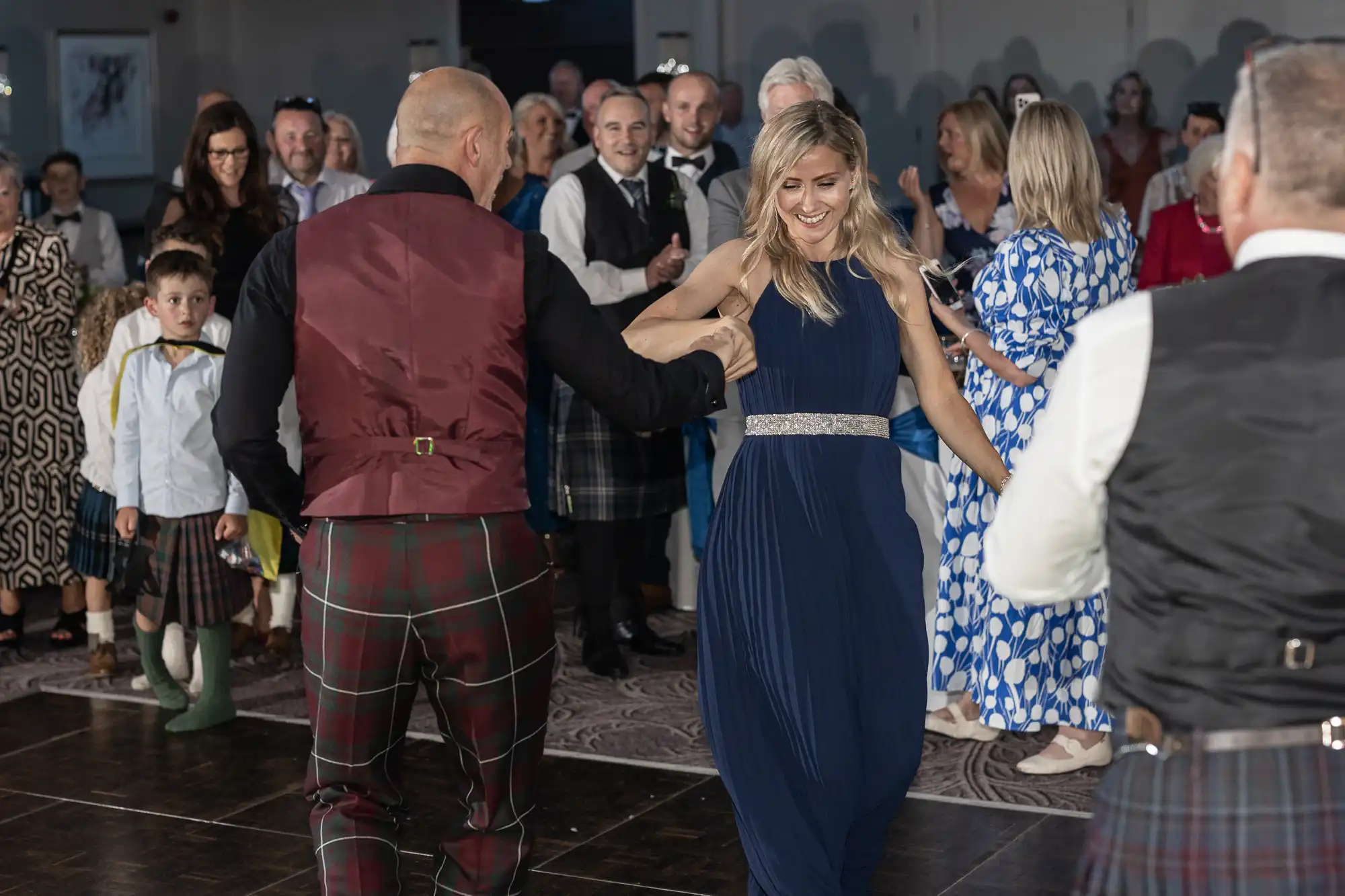 A group of people are dancing together at a gathering. A man and a woman wearing formal attire are dancing in the foreground, with others watching and smiling in the background.