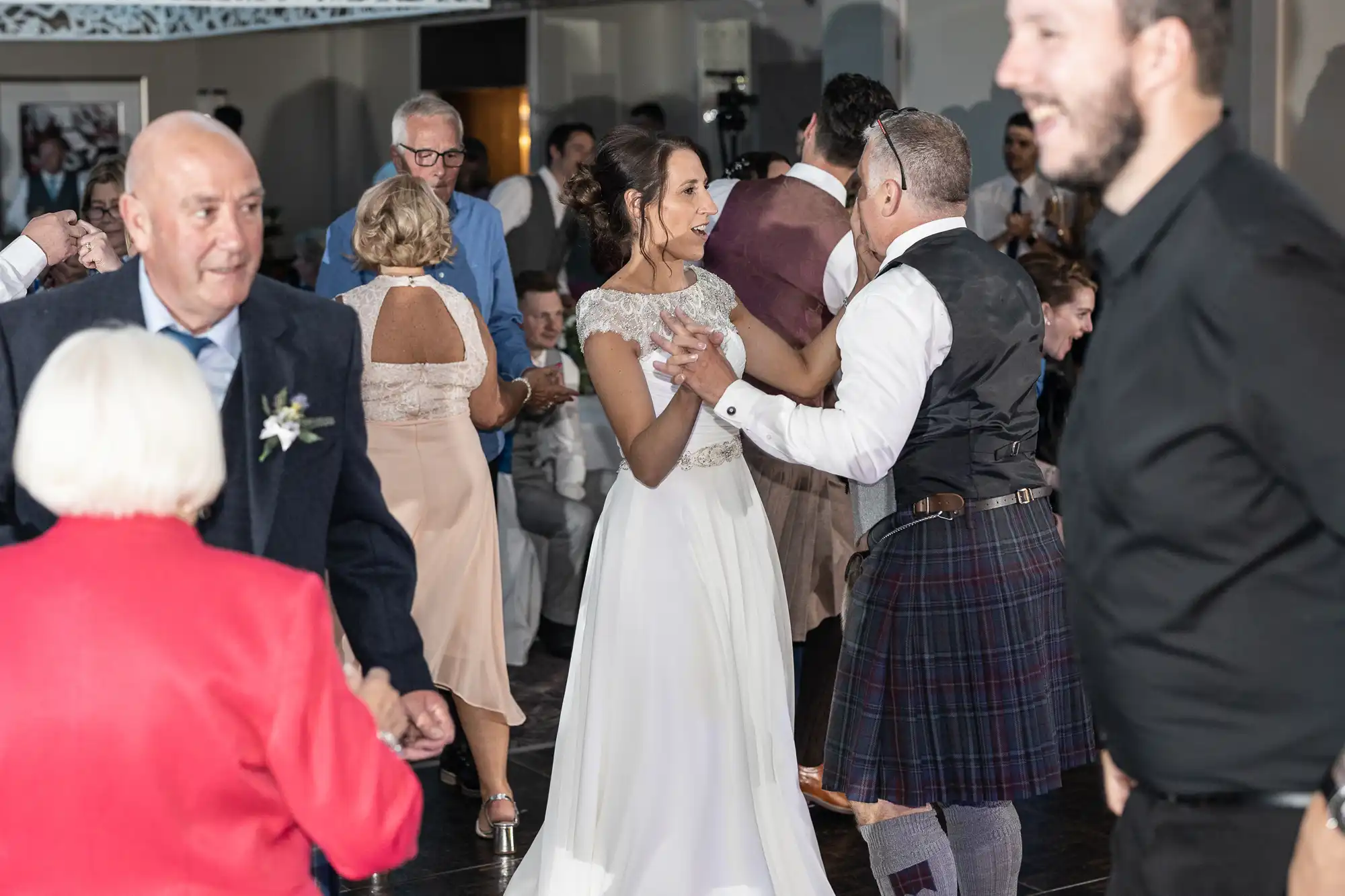 A bride in a white dress dances with a man in a kilt at a lively indoor event, surrounded by dancing guests of various ages.