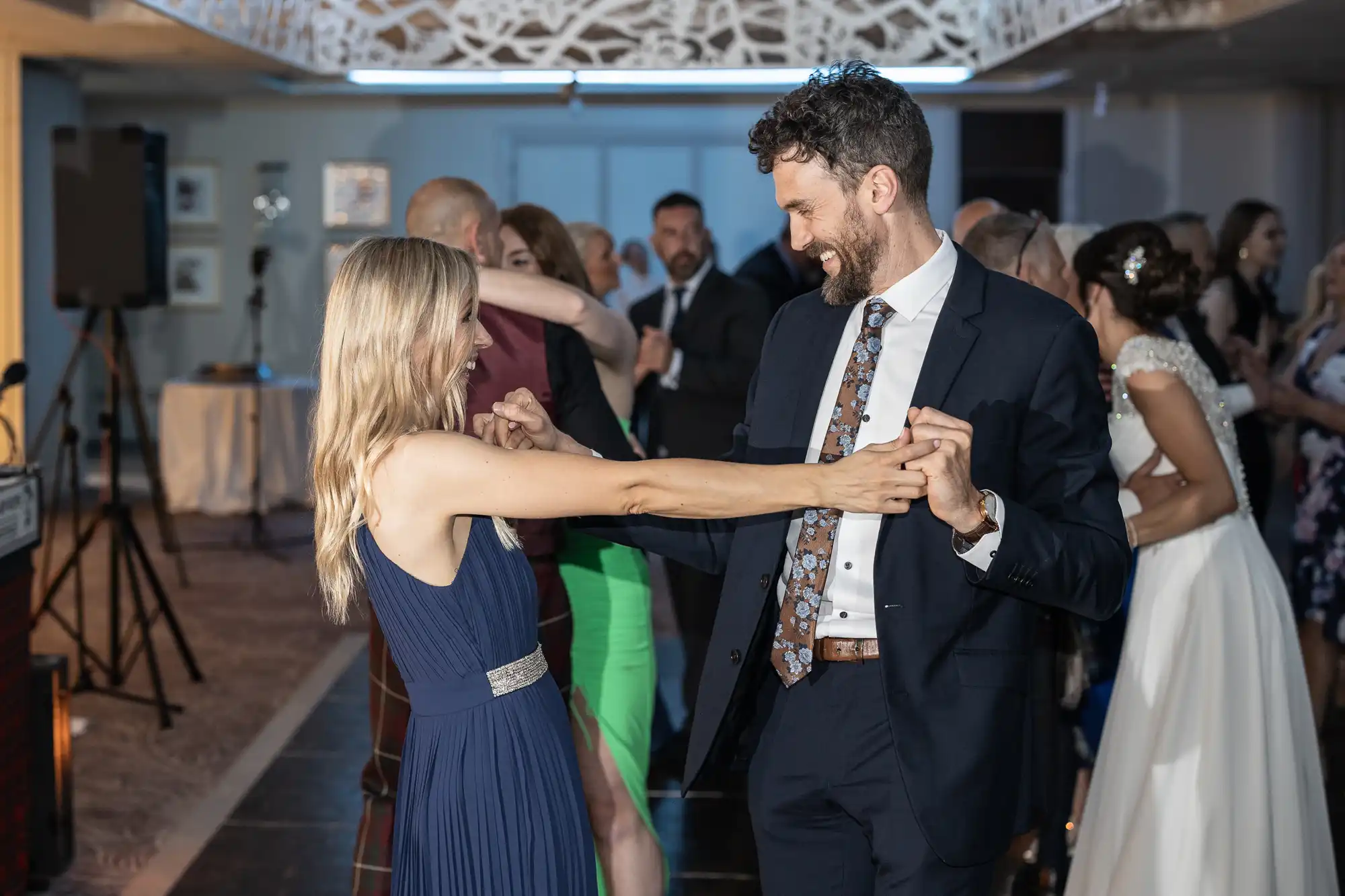 A man and a woman dance together joyfully in a well-lit room with other people socializing in the background.