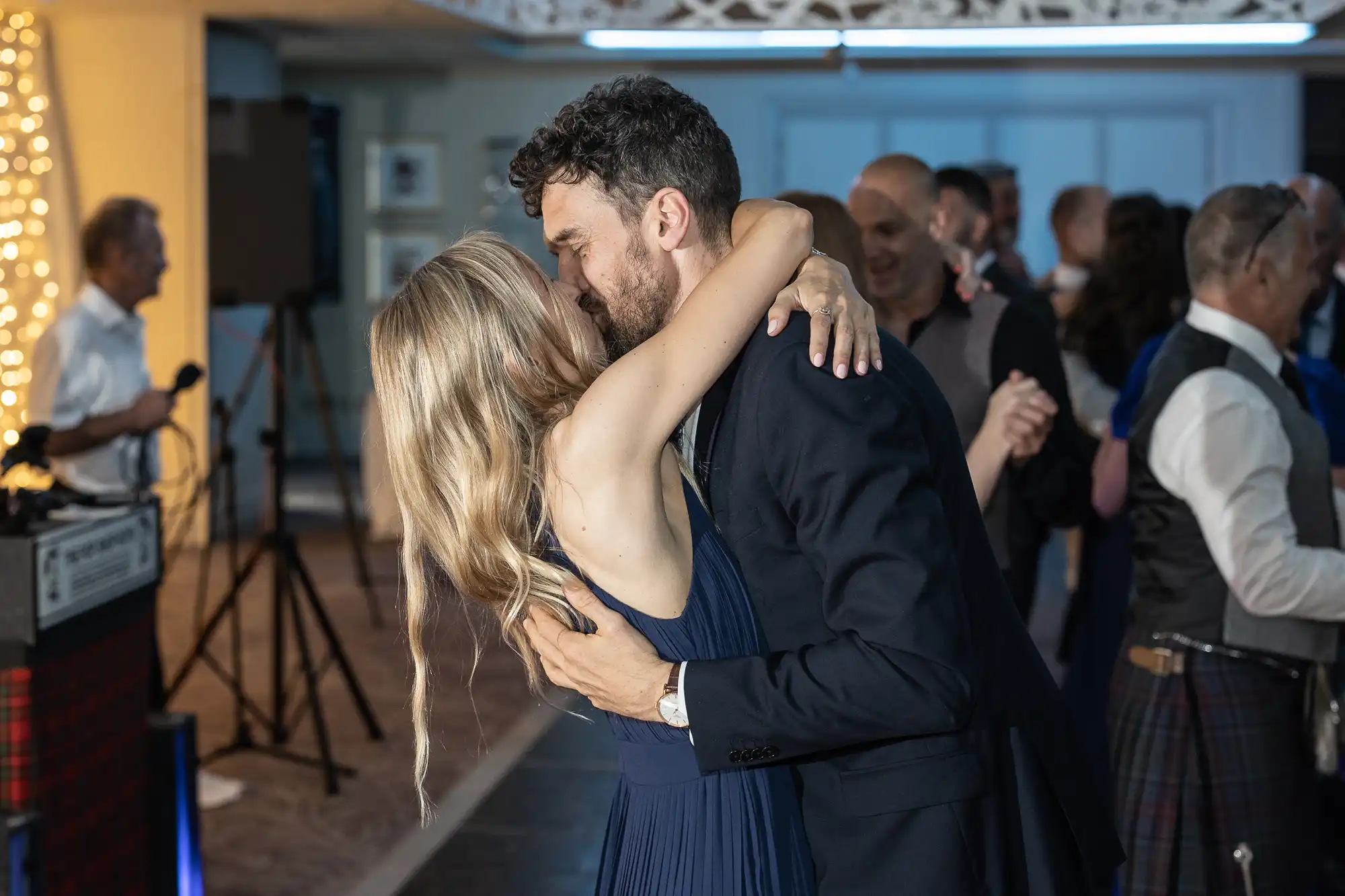 A couple is dancing closely at an indoor event. The man is wearing a dark suit, and the woman is in a blue dress. Other people are mingling in the background.