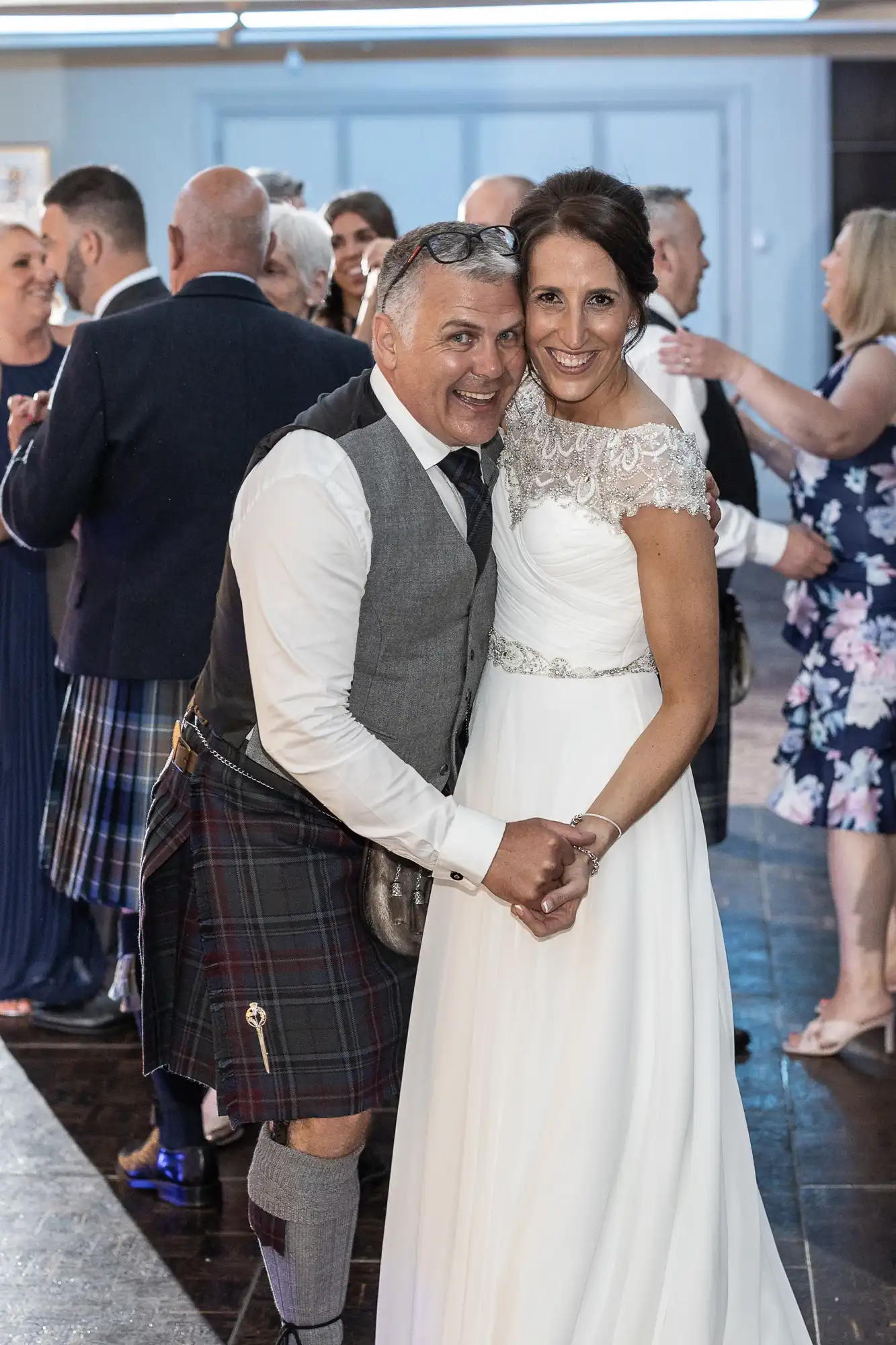 A smiling couple dressed in wedding attire, with the man in a kilt and the woman in a white dress, stand close together while holding hands at a social event with others in the background.