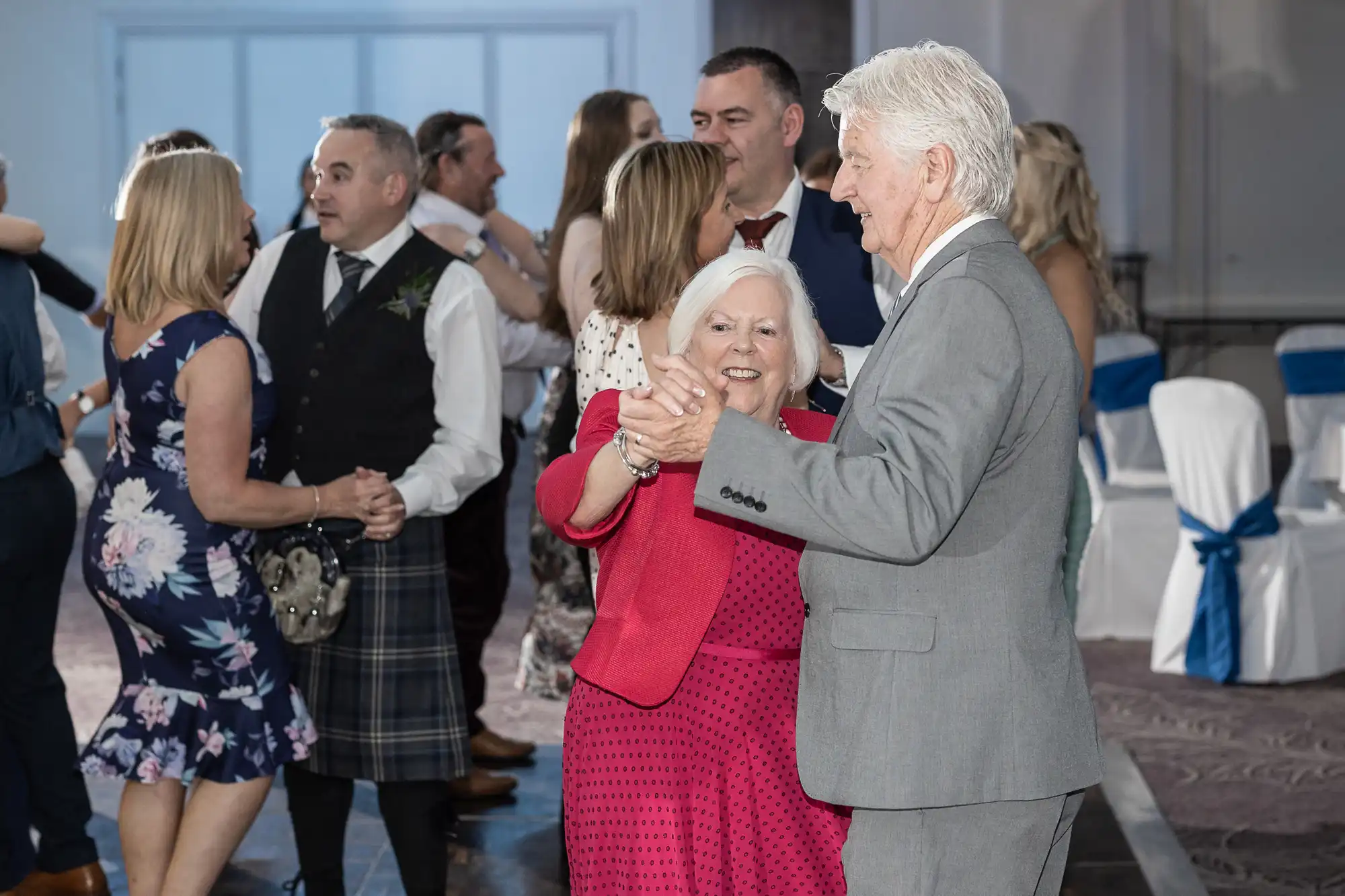 A group of people are dancing at an indoor event. In the foreground, an elderly couple dressed in formal attire dances together, while others are seen dancing in the background.