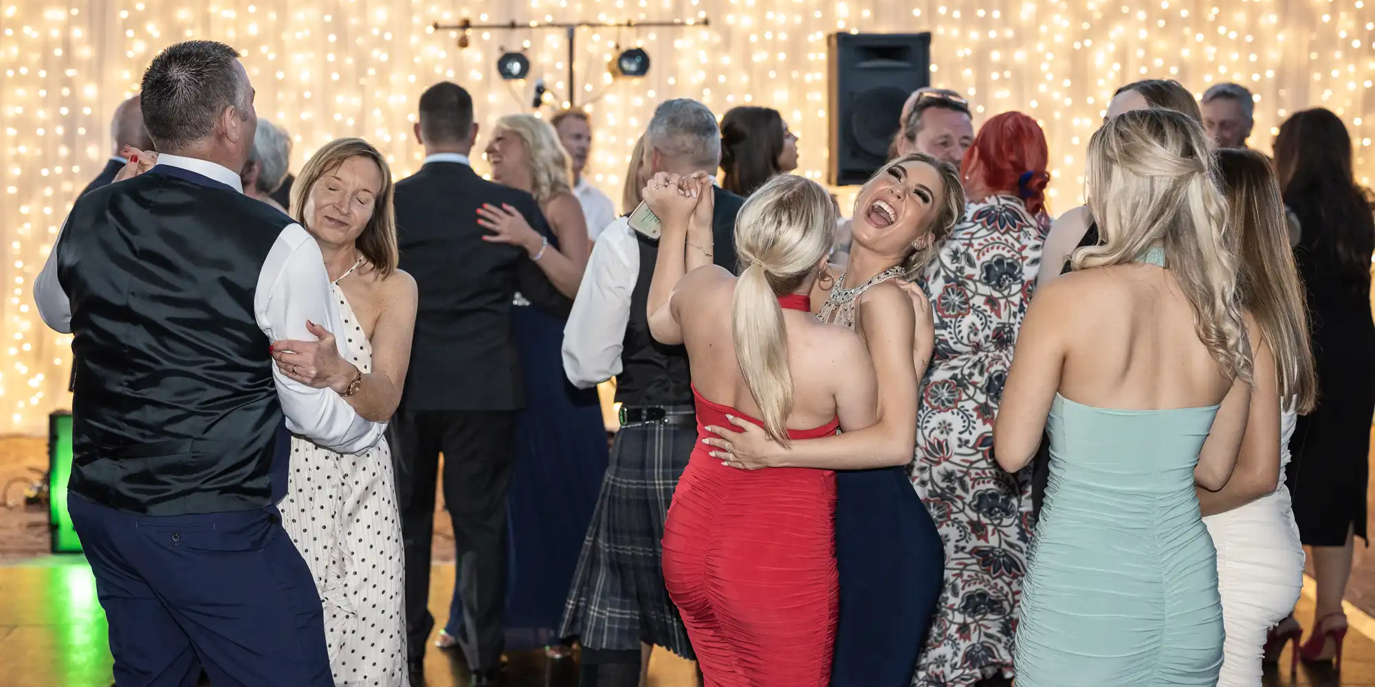 A group of people dancing at a social event, with many couples visible. The background is adorned with string lights, creating a festive atmosphere. Everyone appears to be enjoying themselves.