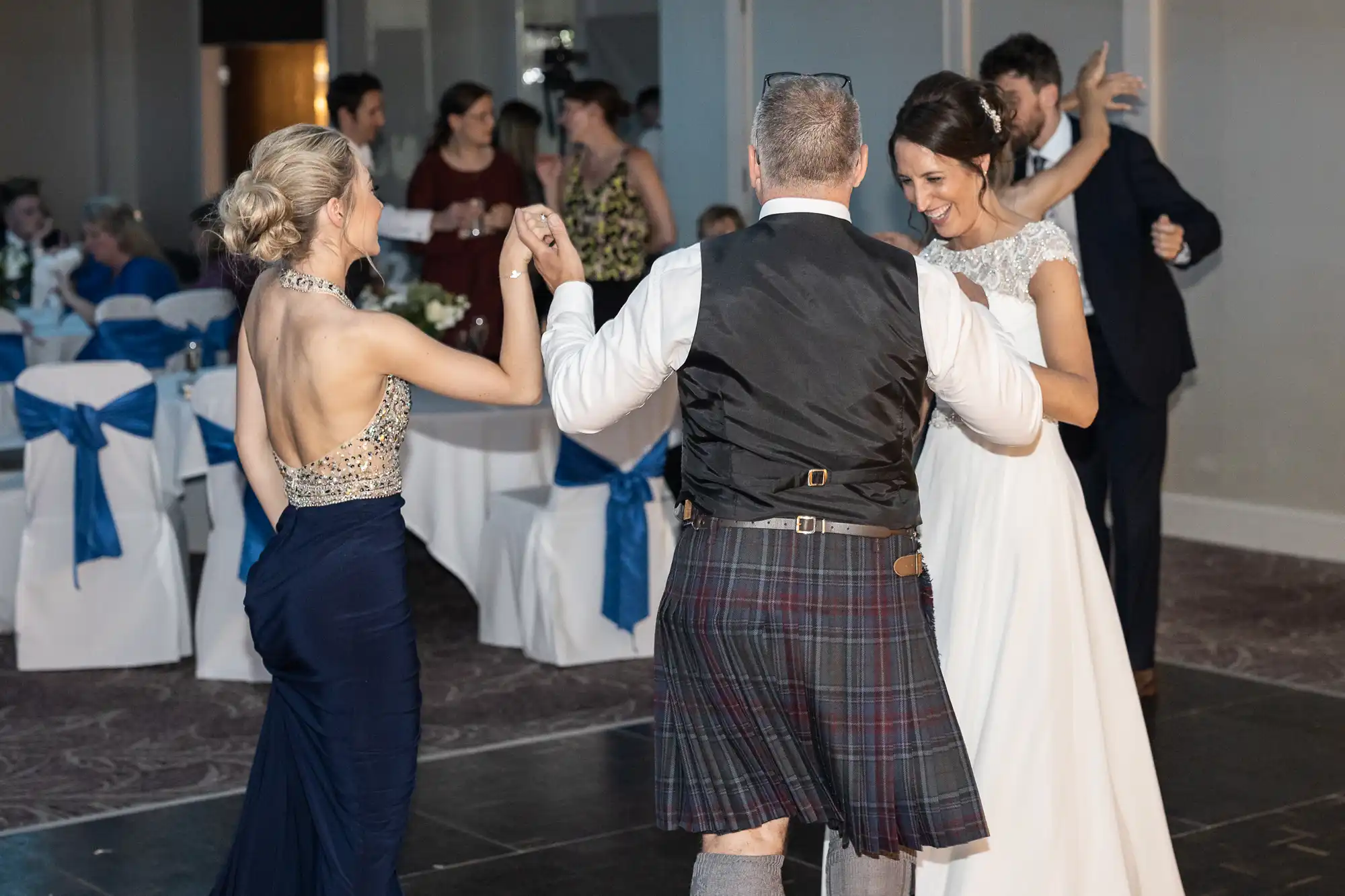 A group of people in formal attire are dancing at an indoor event. A man in a kilt, vest, and shirt is dancing with two women, one in a navy gown and the other in a white dress.