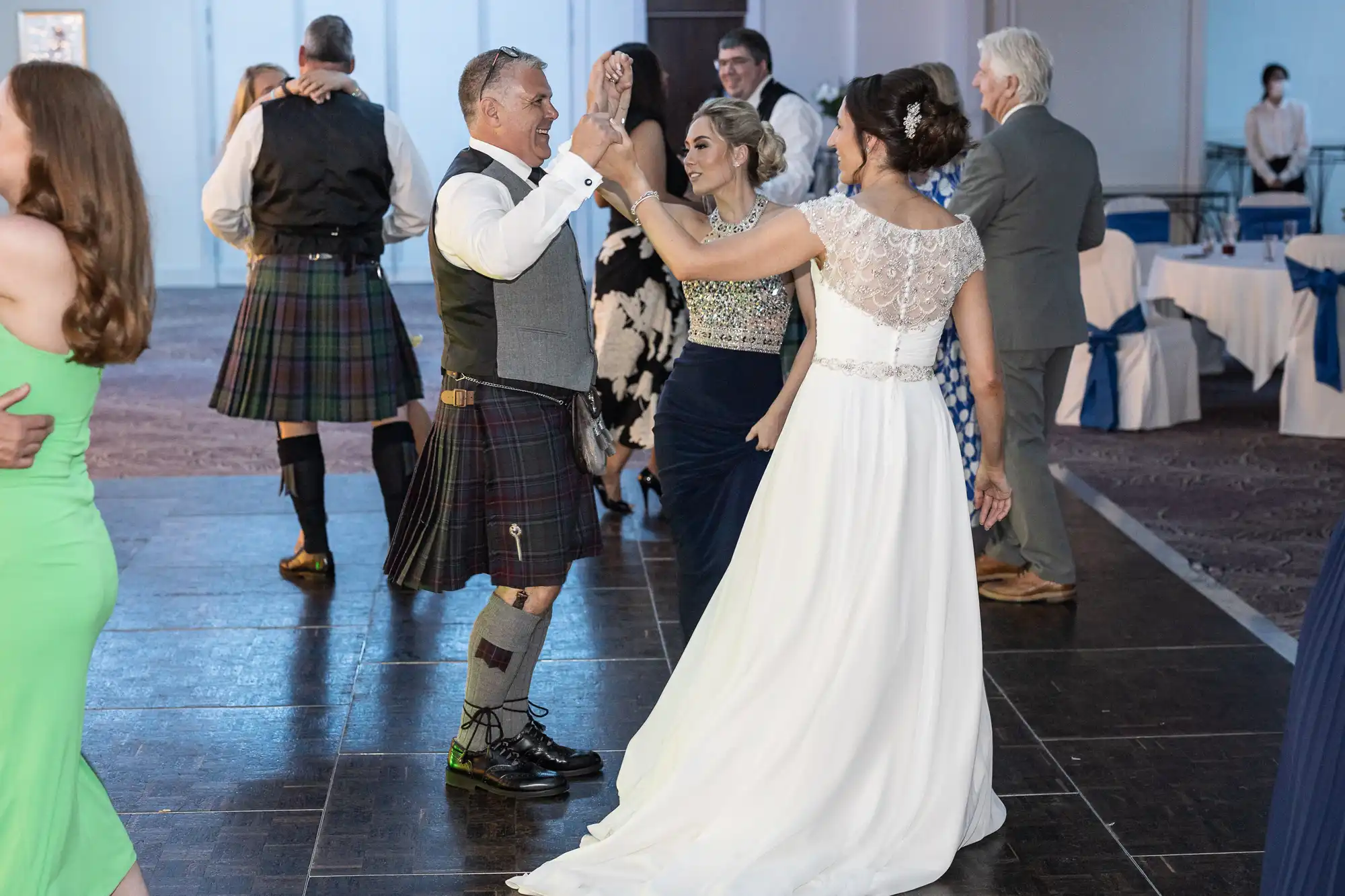 A man in a kilt and a woman in a white dress dance together at an event. Other guests are dancing and mingling in the background.