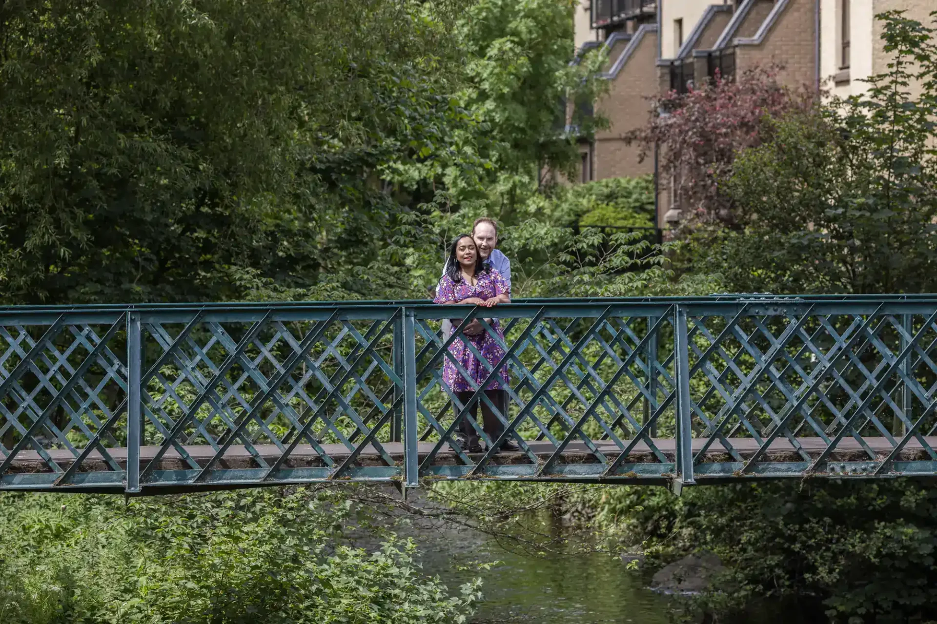 A couple stands together on a metal footbridge surrounded by greenery, with residential buildings visible in the background.