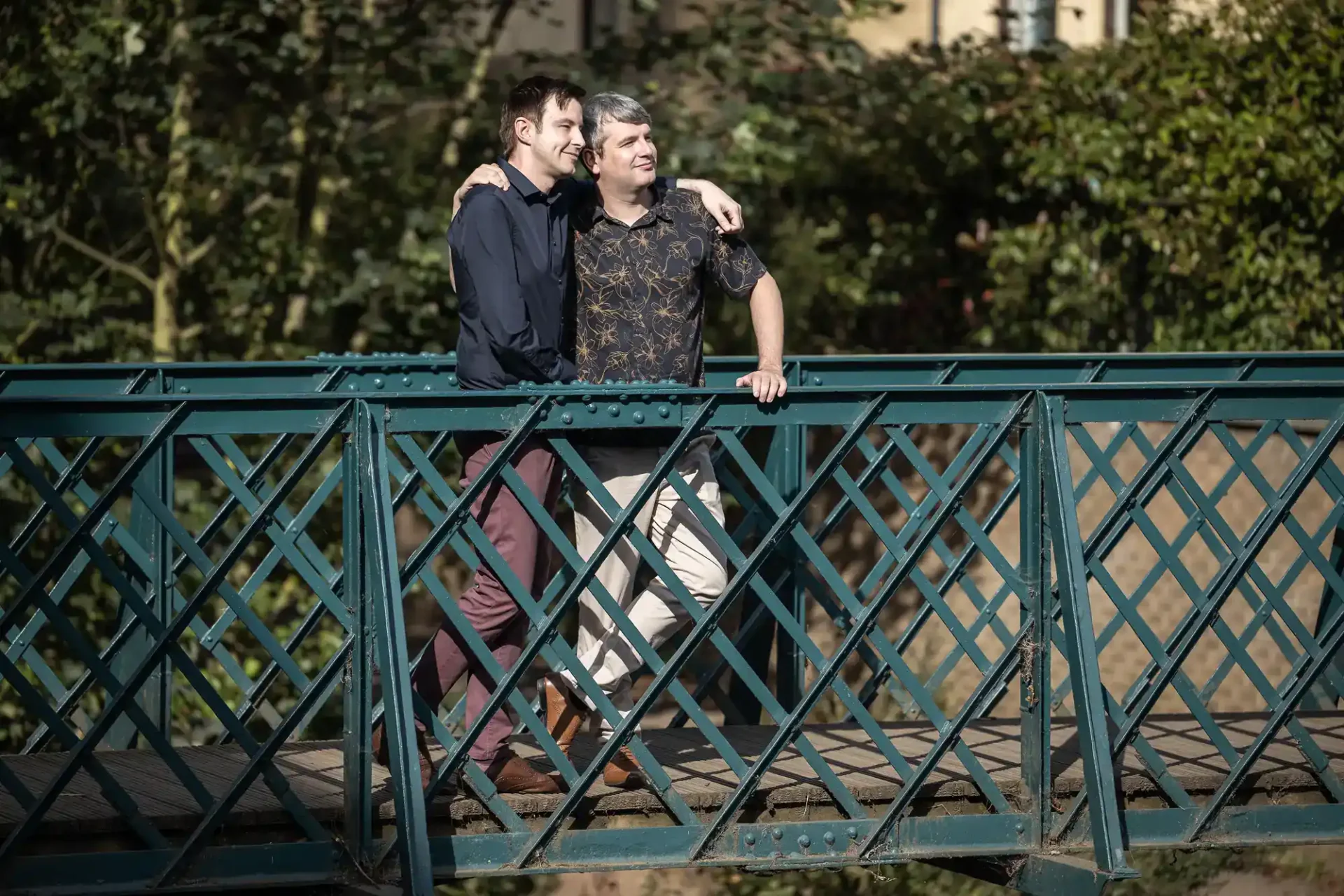 Two men stand side by side, smiling and walking across a green metal bridge, with trees and a building in the background.