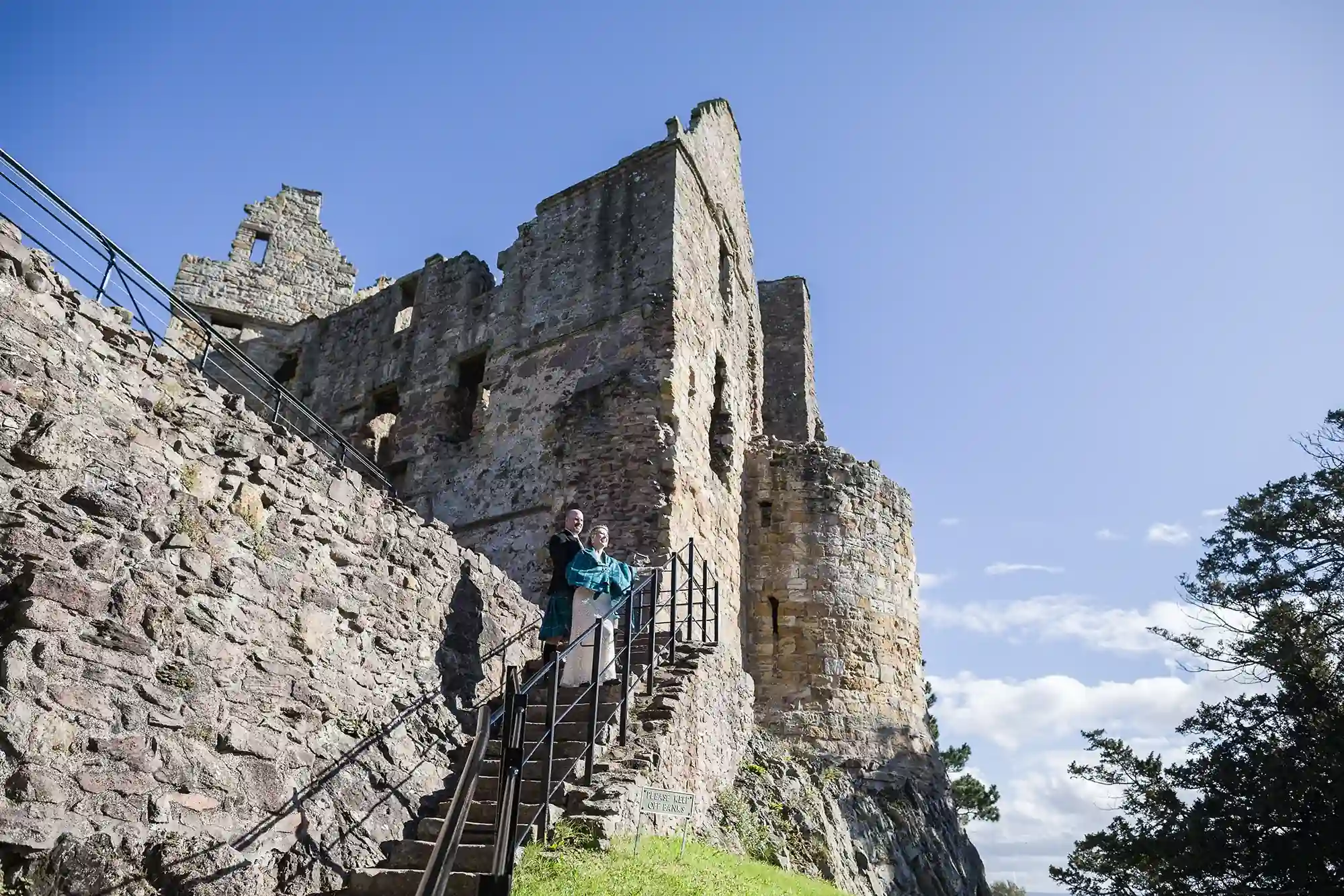 Two people smiling on a staircase beside the ruins of an ancient stone castle under a clear blue sky.