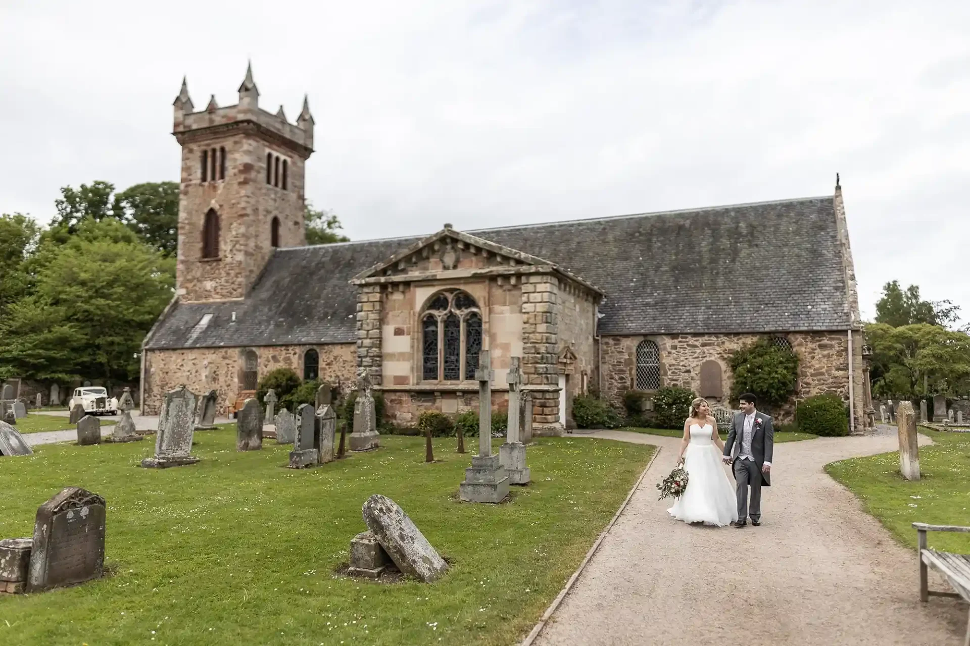 A bride and groom walking hand in hand through a cemetery next to an old stone church on a cloudy day at Dirleton Kirk.