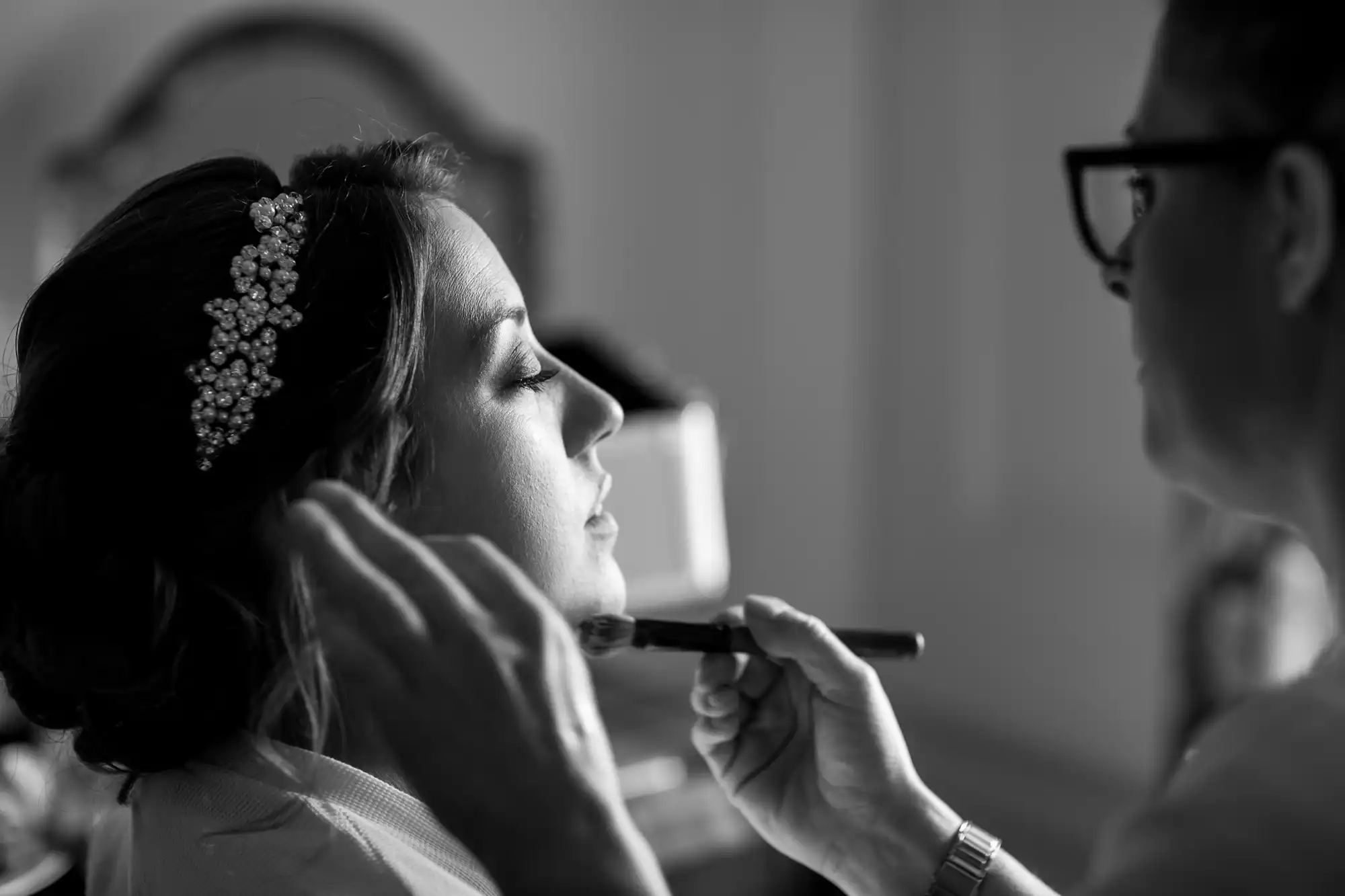 A bride with a beaded hair accessory is having makeup applied by another person holding a brush. The scene is in black and white.