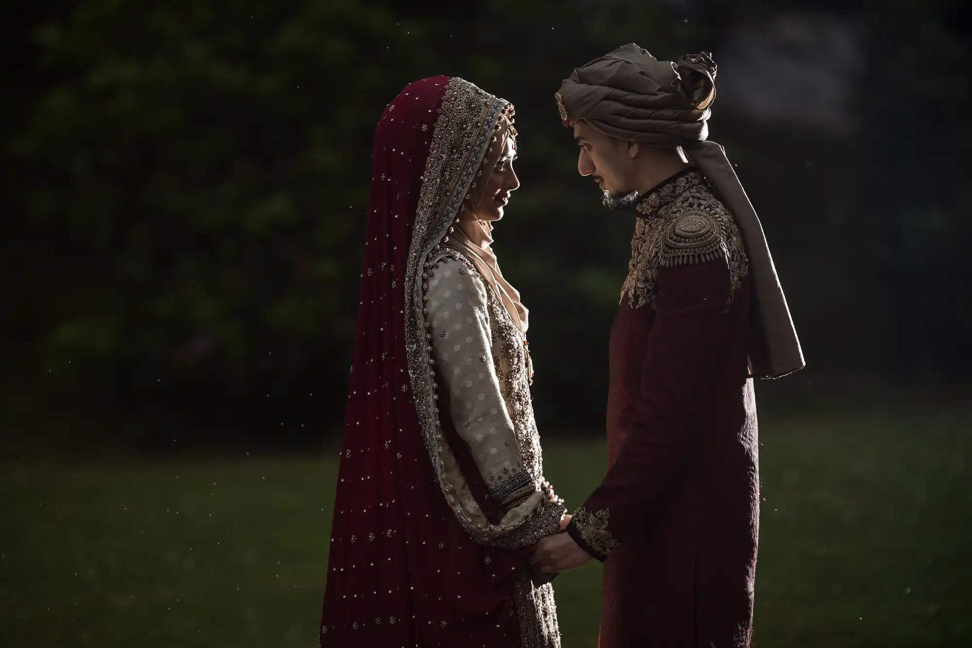 A couple in traditional attire gazes at each other, holding hands in an outdoor setting with a dark, blurred background. The woman wears a red and gold outfit with a veil, and the man wears a matching outfit.
