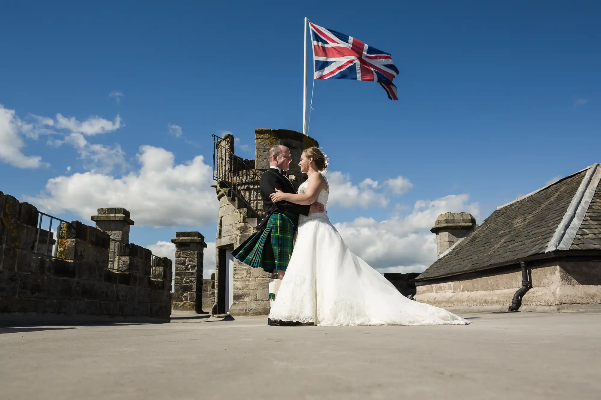 A couple stands on a rooftop under a Union Jack flag; the man wears a kilt and the woman a wedding dress, both in a loving embrace. Stone towers and a blue sky with clouds are in the background.